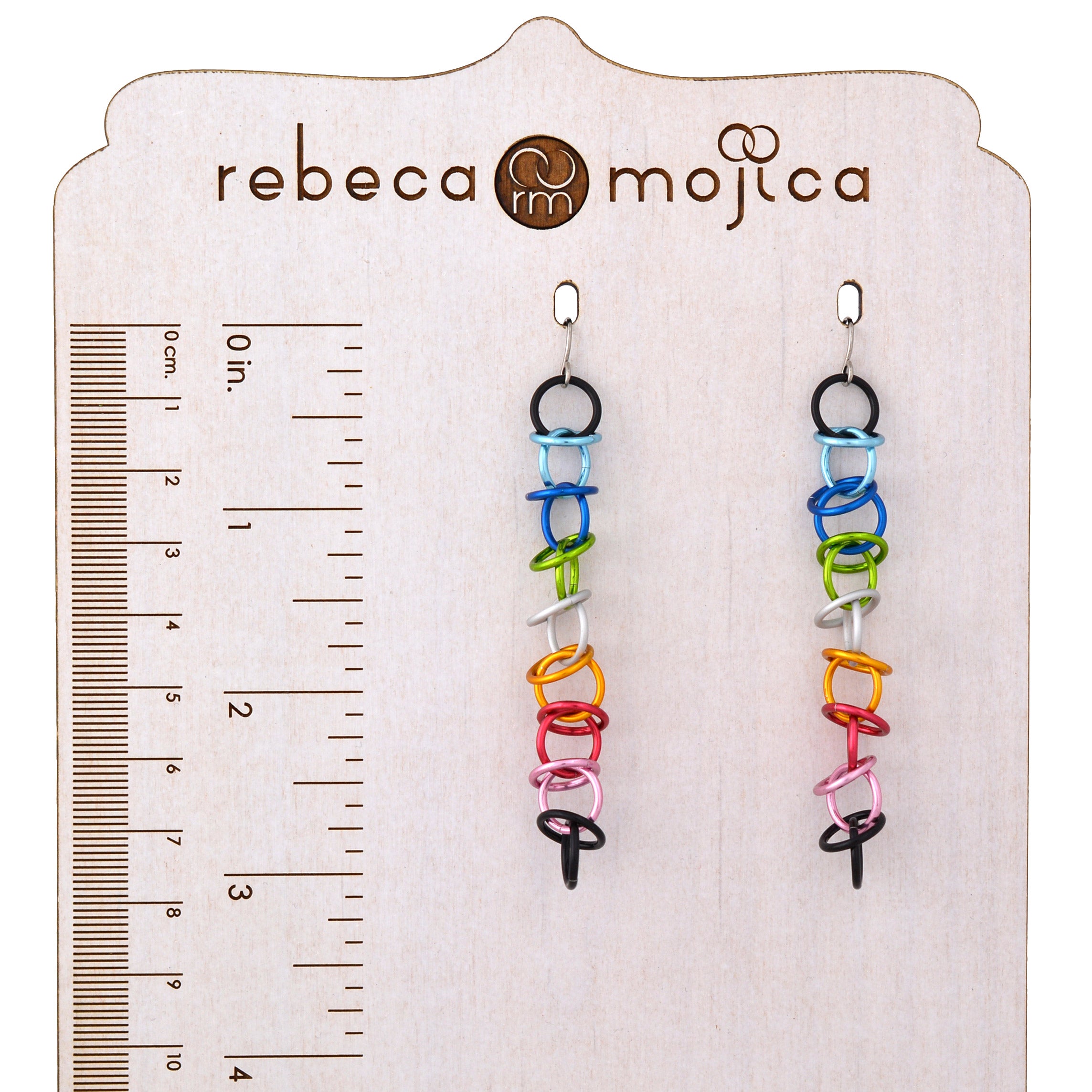 A pair of Queer Pride orbital earrings are displayed on a branded wooden stand, which includes a ruler marked in both centimeters and inches. The earrings are crafted from small, colorful aluminum rings linked together to show the Queer Pride flag colors. The ruler indicates an approximate length of 3.25 inches, and the stand features the "rebeca mojica" logo at the top.