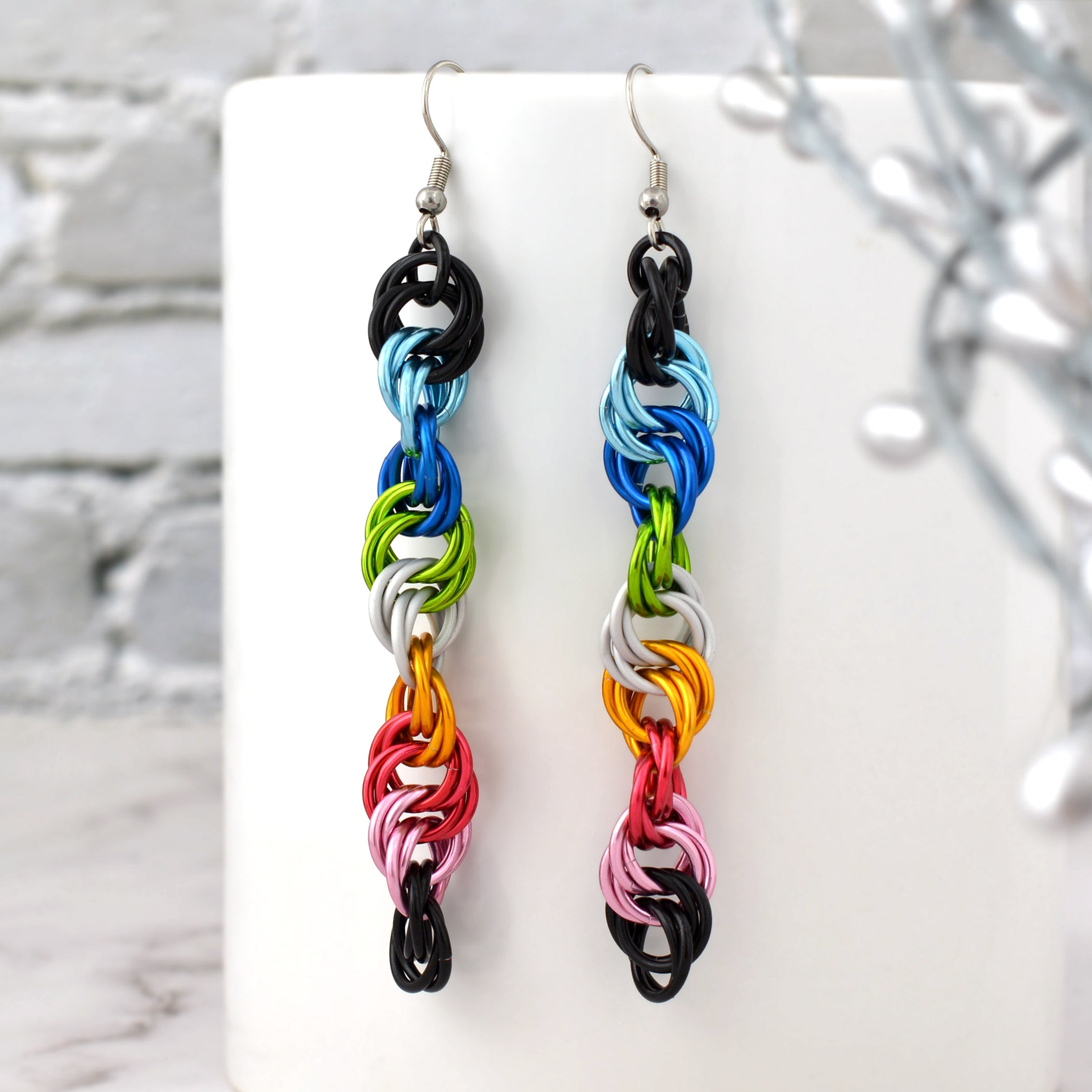 Vibrant queer pride spiral earrings featuring black, light blue, blue, bright green, white, gold, dark pink, light pink rings hang near a blurred white mug and a silver branch detail. Stainless steel hooks. LGBTQ pride flag colors.