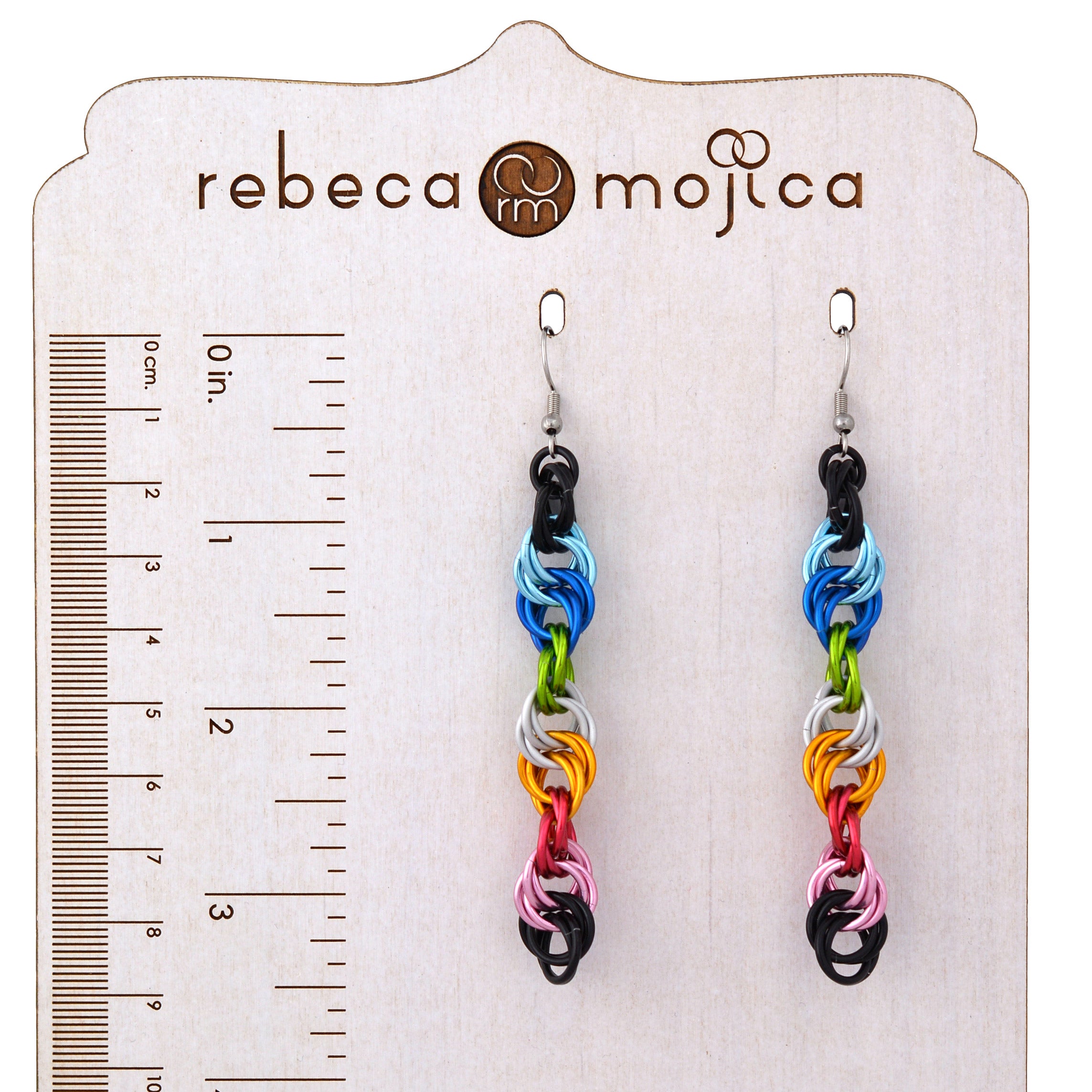 Rebecca Mojica branded earring stand displays long, colorful spiral earrings with a ruler showing cm and inches. Black, light blue, blue, bright green, white, gold, dark pink, light pink rings on silver hooks. "rebeca mojica" logo visible.