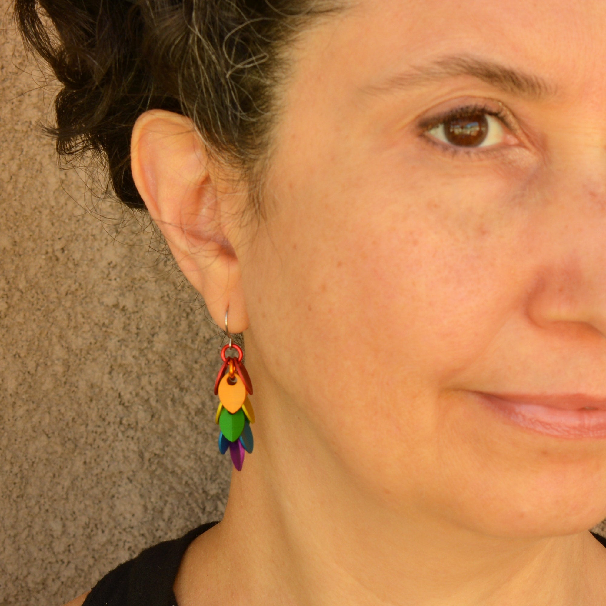 Rebeca Mojica, a person with a slight smile and dark hair, wearing an Aluminum Mini Leaf Pride Dangle Earring from Rebeca Mojica Jewelry, viewed from a slight angle.