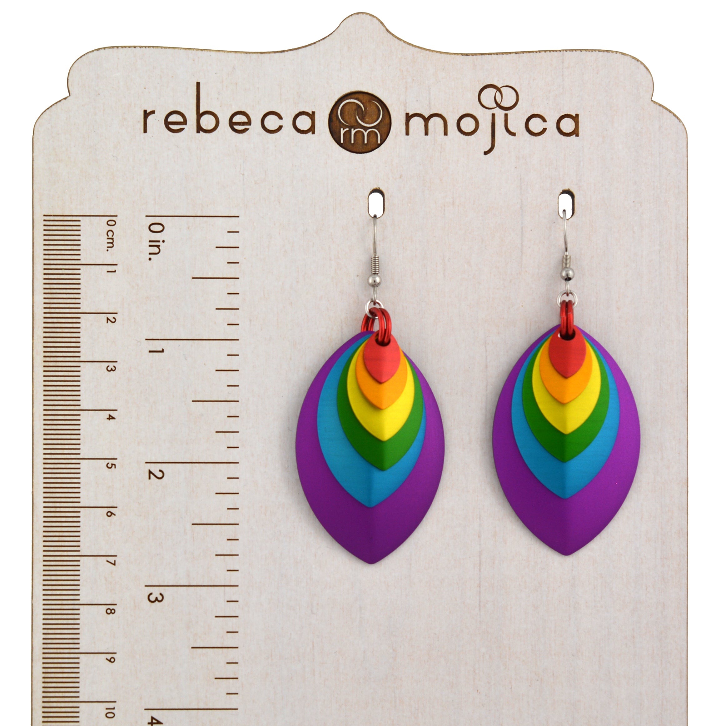 A pair of rainbow-colored stacked leaf earrings with stainless steel fishhook ear wires, placed on a wooden display board with a ruler. The ruler shows the earrings are approximately 2.75 inches long, or about 7 centimeters. The brand name 'rebeca mojica' is visible at the top of the board.