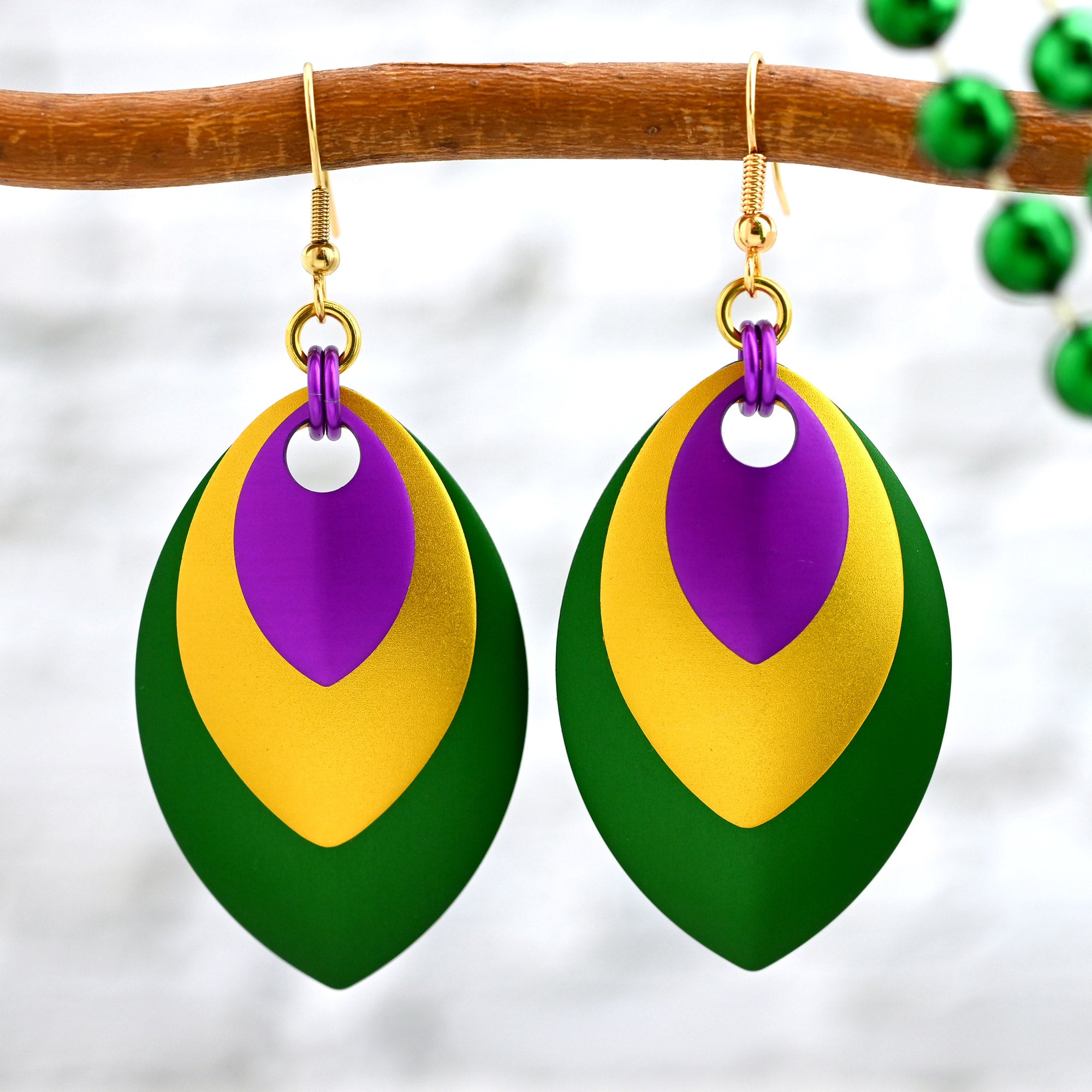 Nested aluminum dragonscale earrings in Mardi Gras colors hanging from a rustic branch with blurred green parade beads