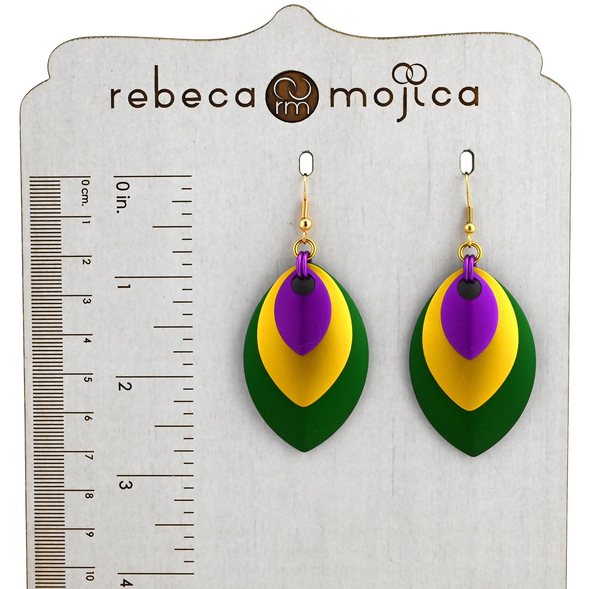Stacked leaf Mardi Gras earrings on a branded Rebeca Mojica display stand with ruler lines showing a length of approximately 2.75 inches.