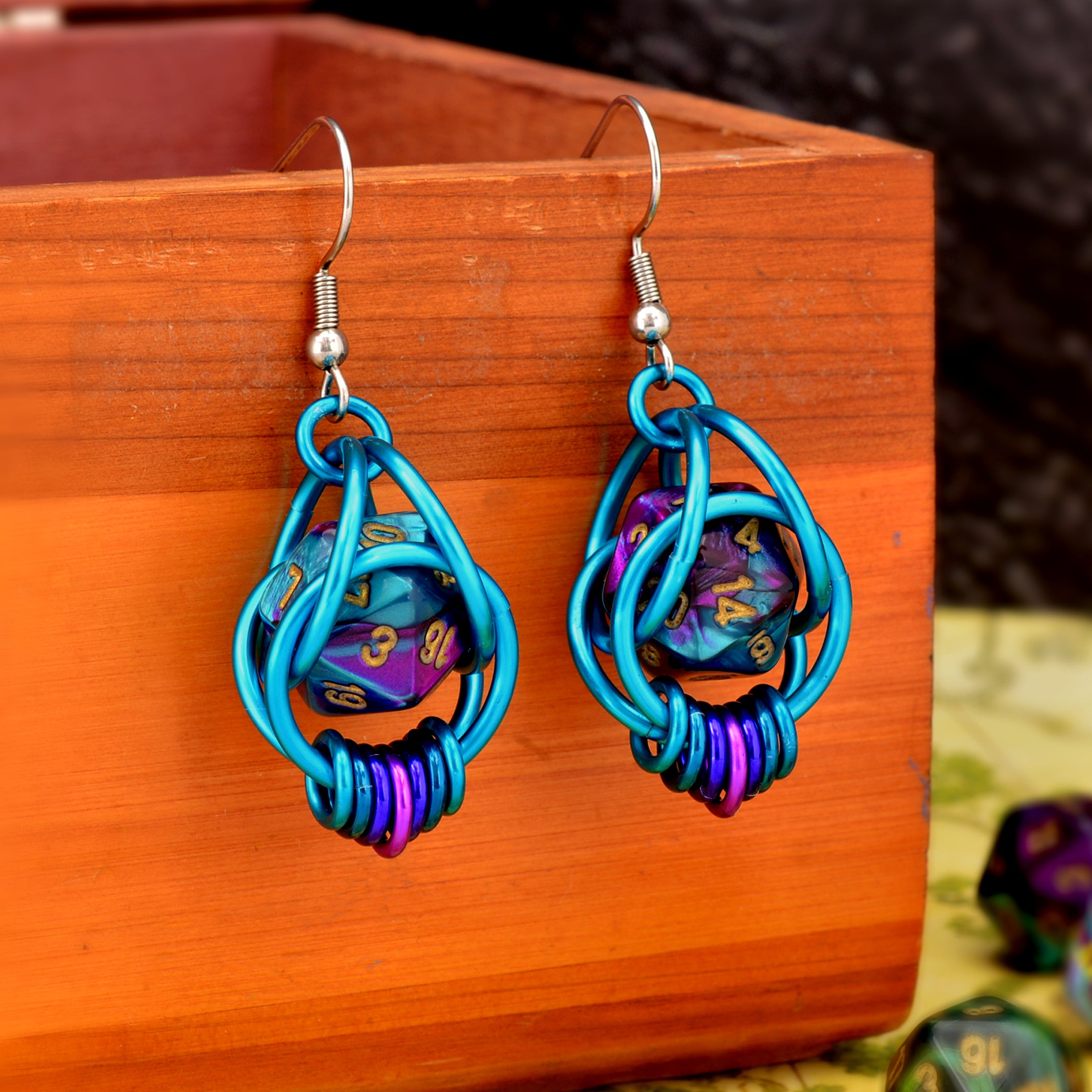 Chainmaille dice earrings featuring mini d20 in marbled purple and turquoise with gold numbers encased in a bright aqua aluminum cage. The earrings are draped over a wooden box with a blurred map and additional loose dice in the background, emphasizing the tabletop gaming theme.