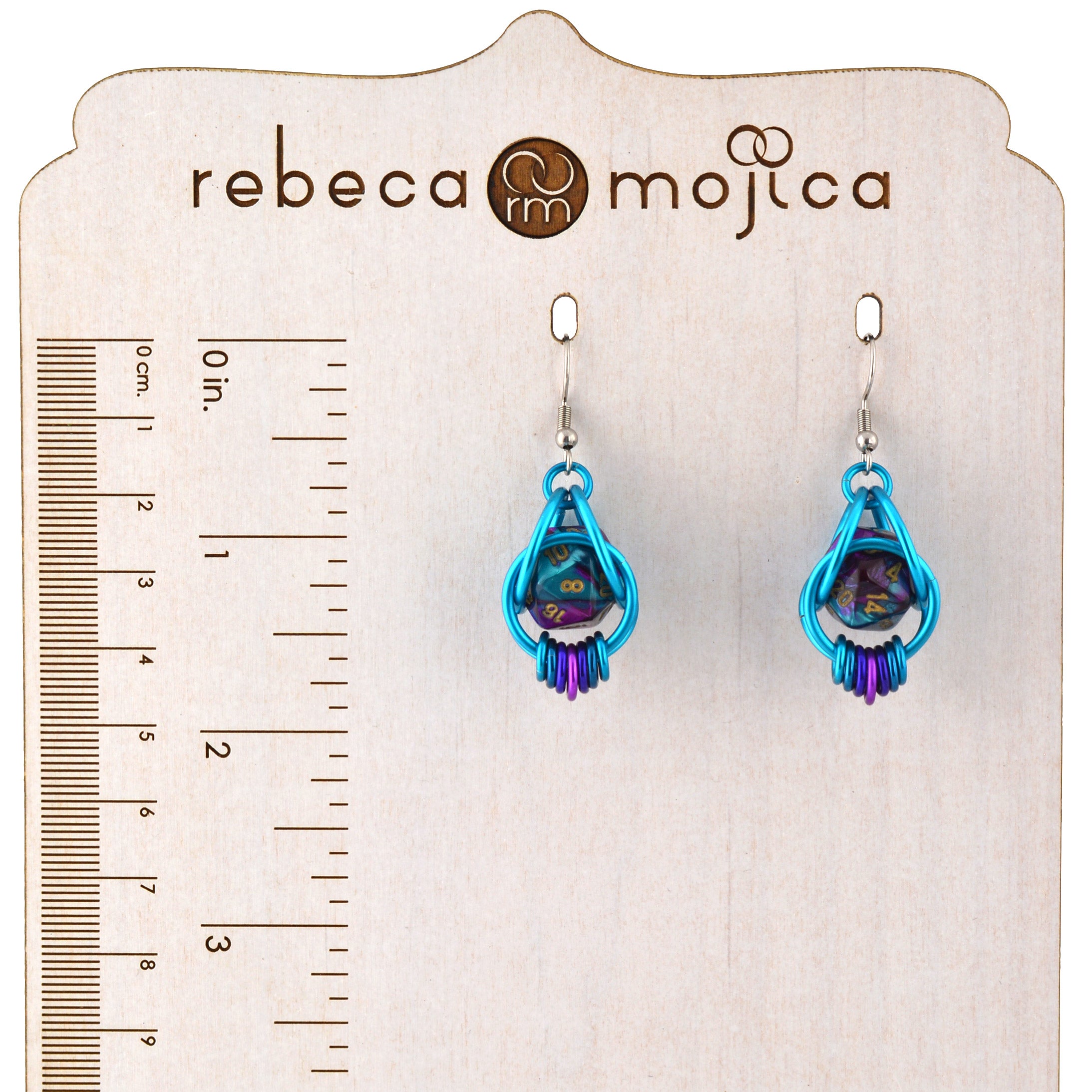 The earrings displayed on a branded "Rebeca Mojica" wooden card next to a ruler, showing the total length is approximately 1.75 inches (4.5 cm).
