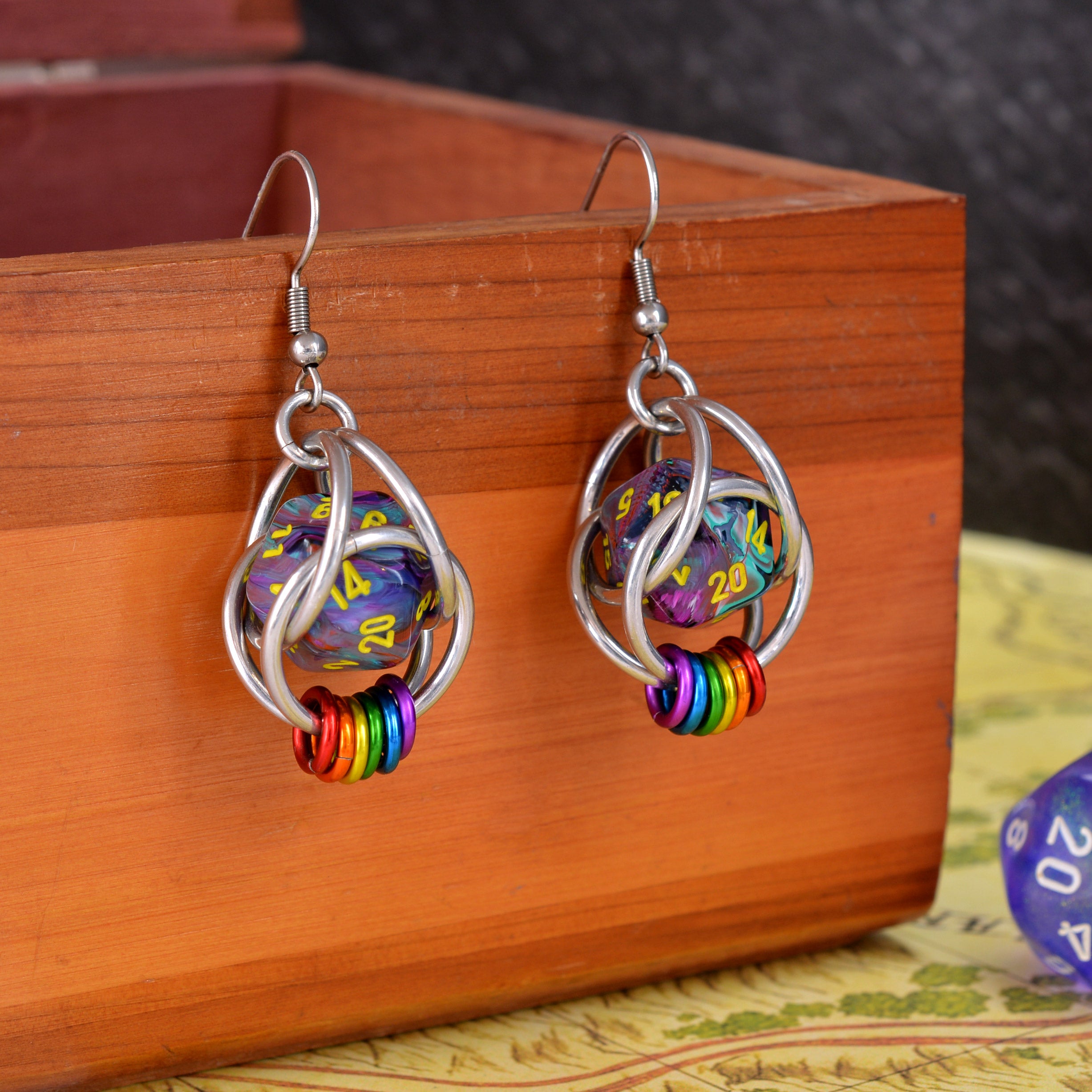 Chainmaille d20 earrings by Rebeca Mojica hanging on a wooden box. Each earring has a mini multi-colored swirl 20-sided die with yellow numbers, encased in a 4-ring aluminum cage, with 6 tiny rings hanging from the bottom in the colors of the lgbtq rainbow pride flag.