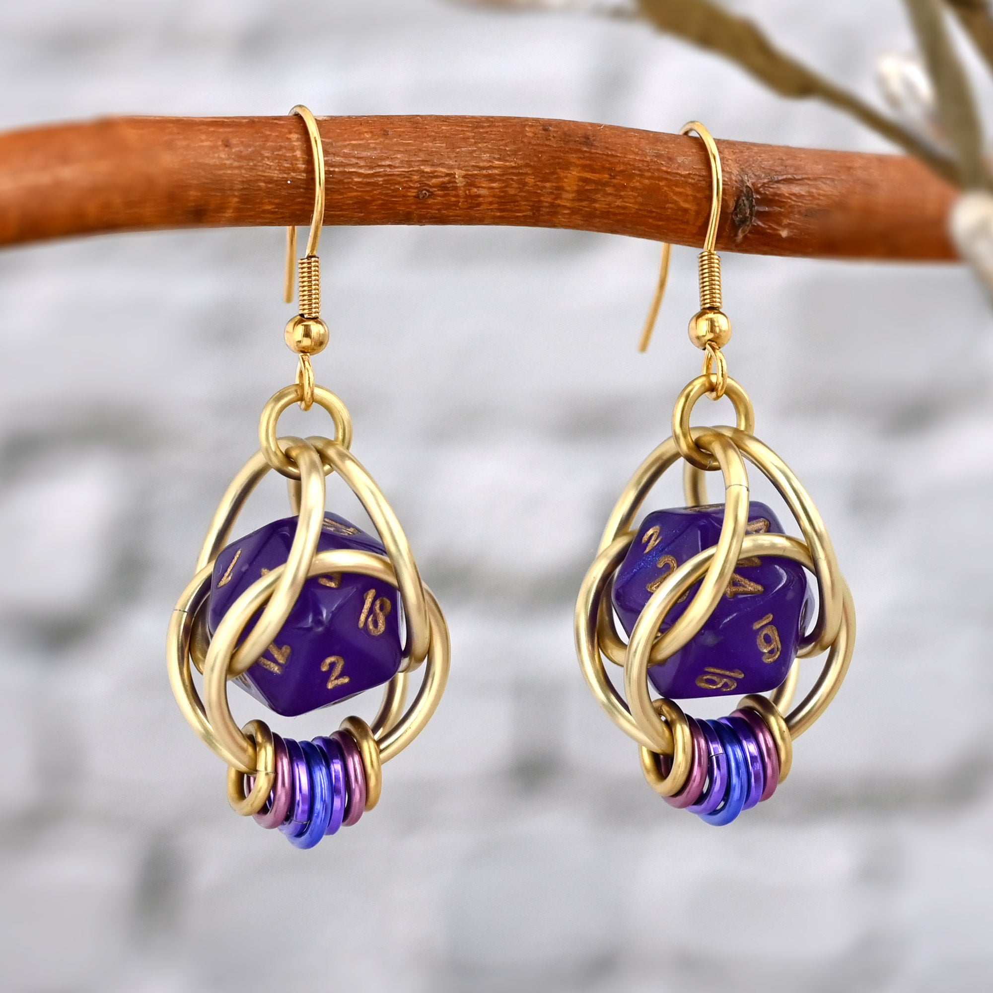 A pair of Wisteria Fae d20 earrings hanging from a natural wooden branch against a light grey brick background. Each earring features a translucent amethyst purple 20-sided die held within a sculptural, hand-linked cage of straw-colored aluminum rings. The base is accented with a decorative fidget coil ombre in shades of violet and blue, creating a whimsical, fantasy-inspired aesthetic.