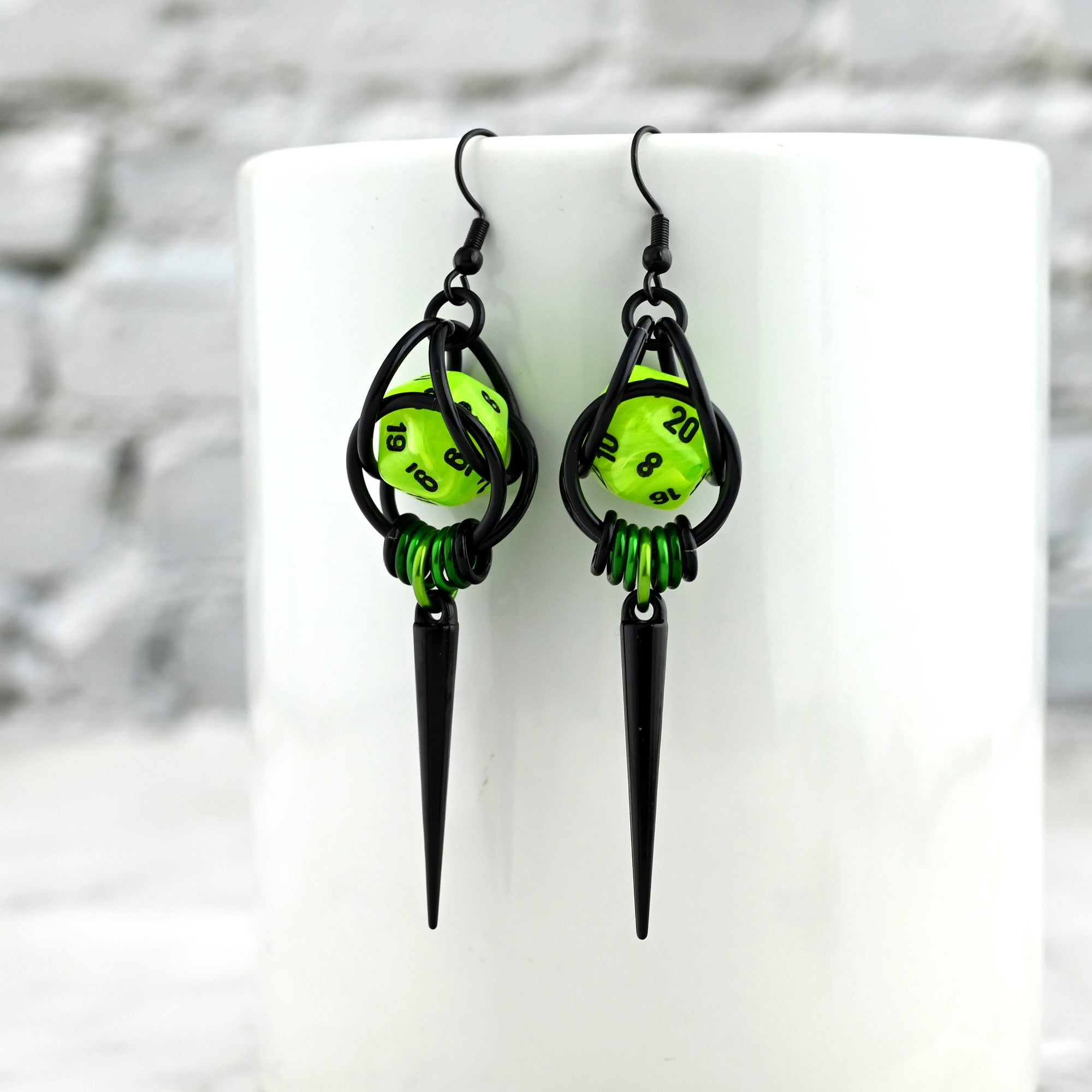Geeky D20 spike earrings hanging on a white ceramic mug. The bright neon green of the marbled dice pops against the white surface.