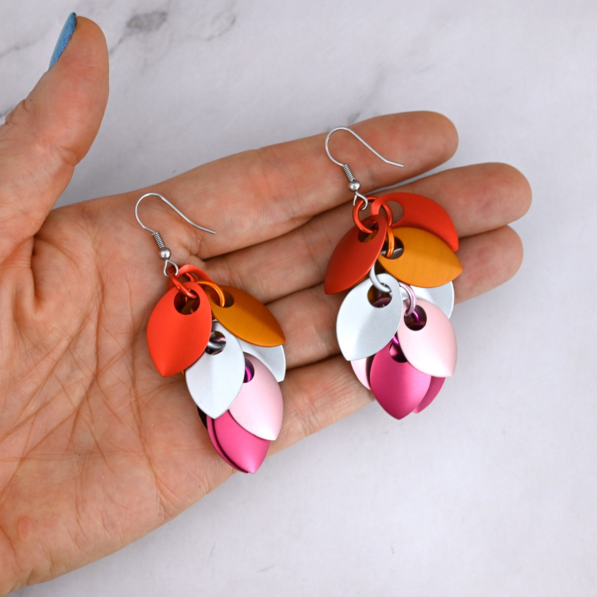 A pair of Lesbian Pride scale earrings held in a person's palm, demonstrating the physical size and the vibrant, matte satin finish of the aluminum leaves.