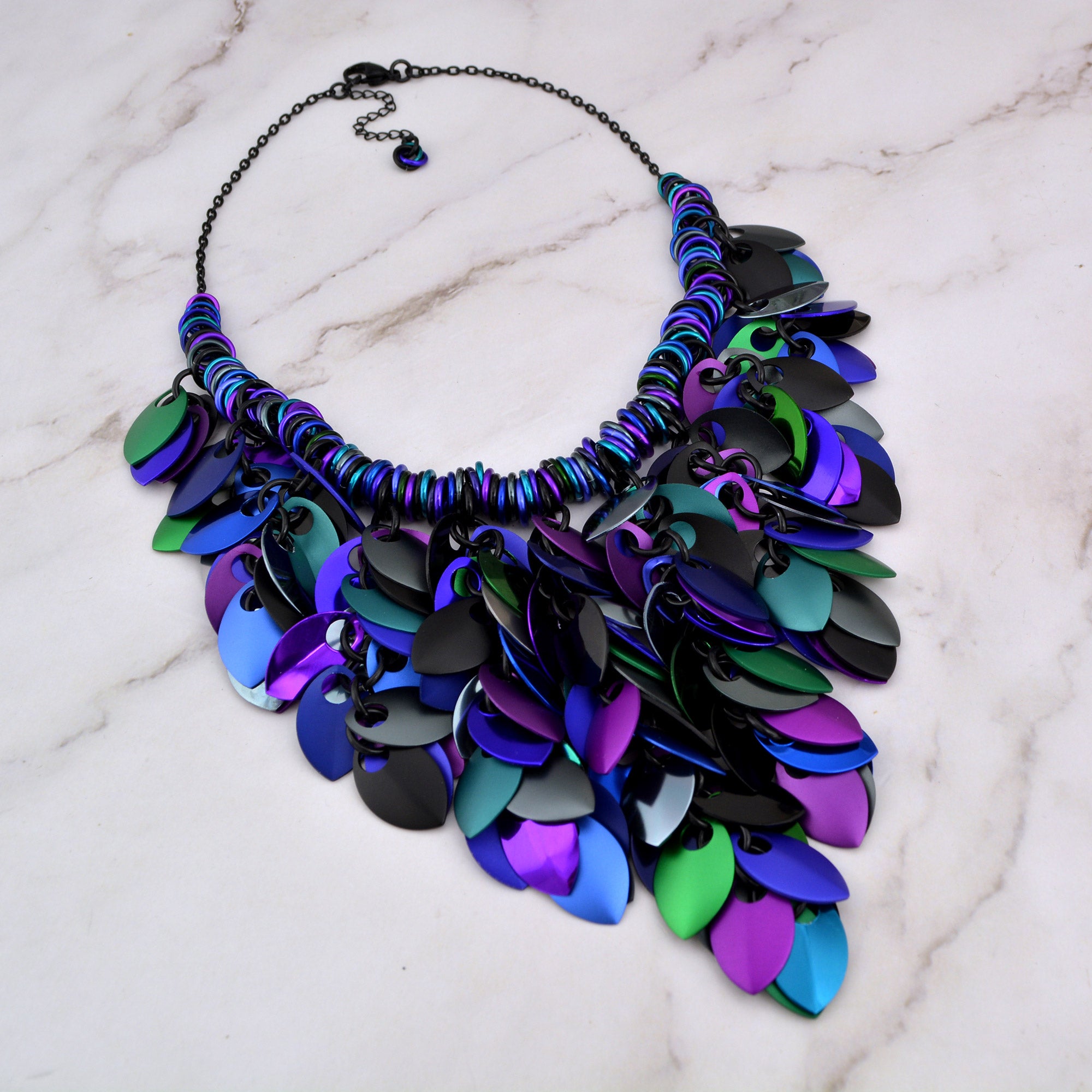 Aerial view of Dark Mermaid scalemaille necklace on a marble background, showcasing the dark and colorful scales in front, and the black chain, clasp and extender in rear.