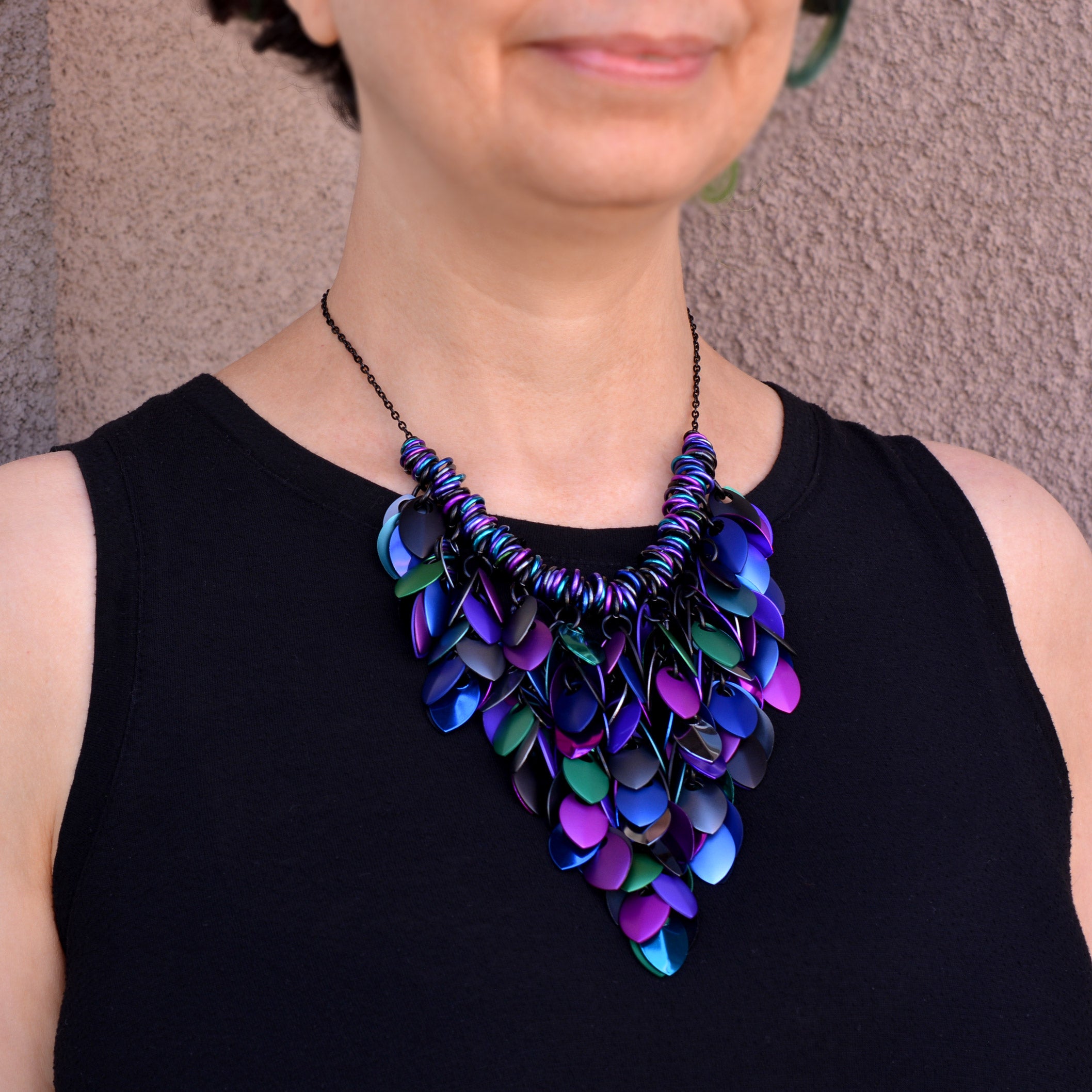 A medium shot of a person wearing the Dark Mermaid Feathered Bib necklace. The statement scalemaille piece rests prominently on the chest, showcasing its 5-inch height and fluid, "feathered" appearance. The necklace is composed of a dense cluster of colorful metal leaves that shift between matte and reflective finishes. It is attached to a delicate black chain, and the vibrant blues and purples of the jewelry pop against the wearer's black sleeveless shirt.