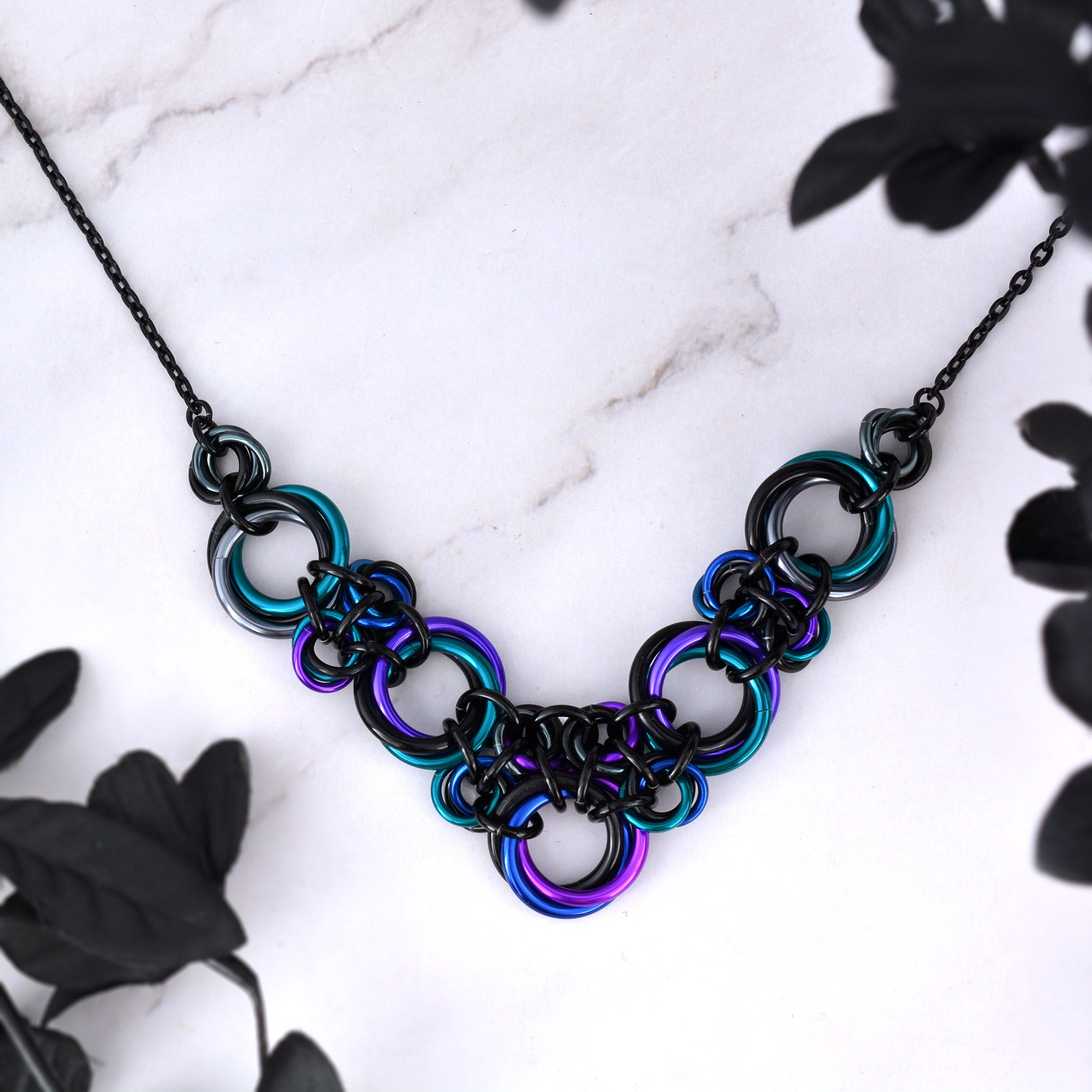 Stylized flatly of Knotted Metal V necklace in Dark Mermaid colors (teal, purple, blue, violet, black, grey), highlighting the chainmaile knot pattern.