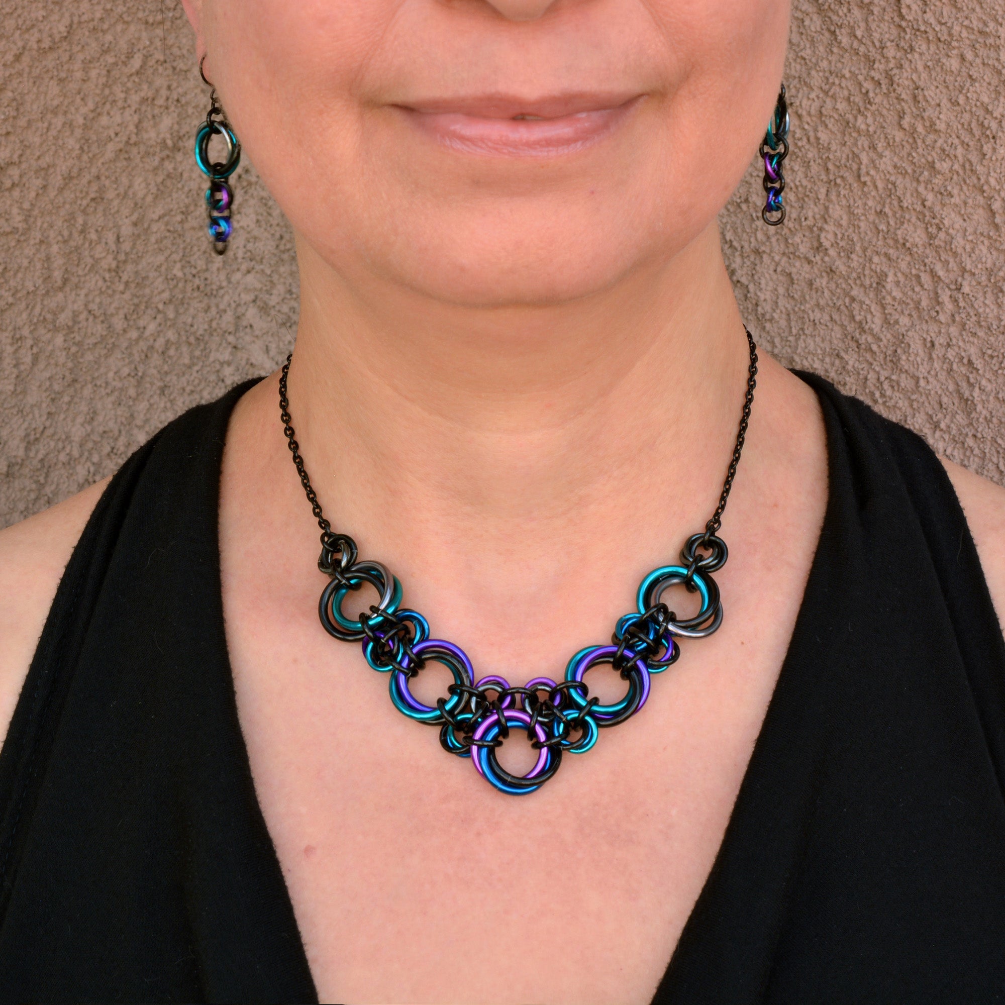 Close-up of Rebeca Mojica wearing the lightweight Knotted Metal V chainmail necklace and Comet earrings in Dark Mermaid (black, teal, purple, blue, violet).