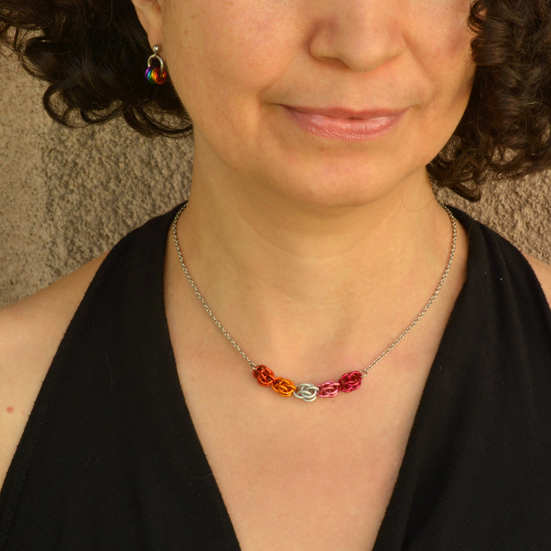 Closeup of short chainmaille necklace being worn. Necklace has a 2-inch focal section attached to a thin steel chain on each end. The focal is made of 6 chainmaille “beads” in the Sweet Pea weave - each segment has 6 links of the same color. From left to right, colors are papaya, orange, white, pink and fuchsia - to best match the lesbian pride flag.