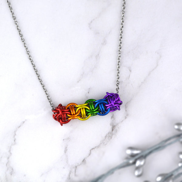Rainbow pride chainmaille sliding bar necklace displayed on a light marble surface. The chainmaille links form a small flexible tube (in the Captive Inverted Round pattern), about 1.5 inches long, that slides along the thin steel chain. Three links of a single color make a triangle that surrounds 2 of the same color link. There is one triangle for each color of the flag: red, orange, yellow, green, blue and purple, and the triangles are joined to each other. Made by Rebeca Mojica in California, USA