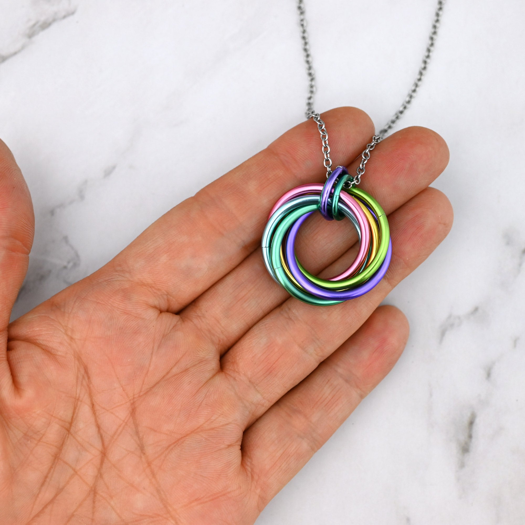 A person holding the Blossom Swirl Chainmaille Pendant in their palm to show scale. The necklace features intertwined light blue, pink, lilac, and lime green aluminum rings on a stainless steel chain