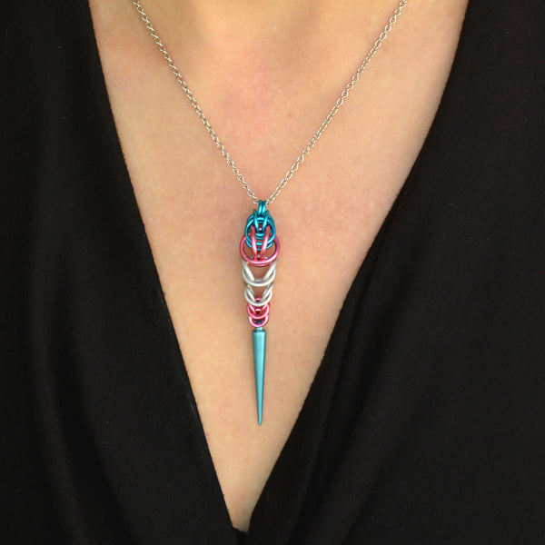 Close-up of a chainmaille pendant on a thin silver chain on a woman’s neck. The pendant is long and narrow, made in the colors of the transgender pride flag. The chainmaille weave is aqua at top, transitioning to pink, white, and then pink again, with a light turquoise acrylic spike at the bottom. The woman is wearing a black V-neck top.