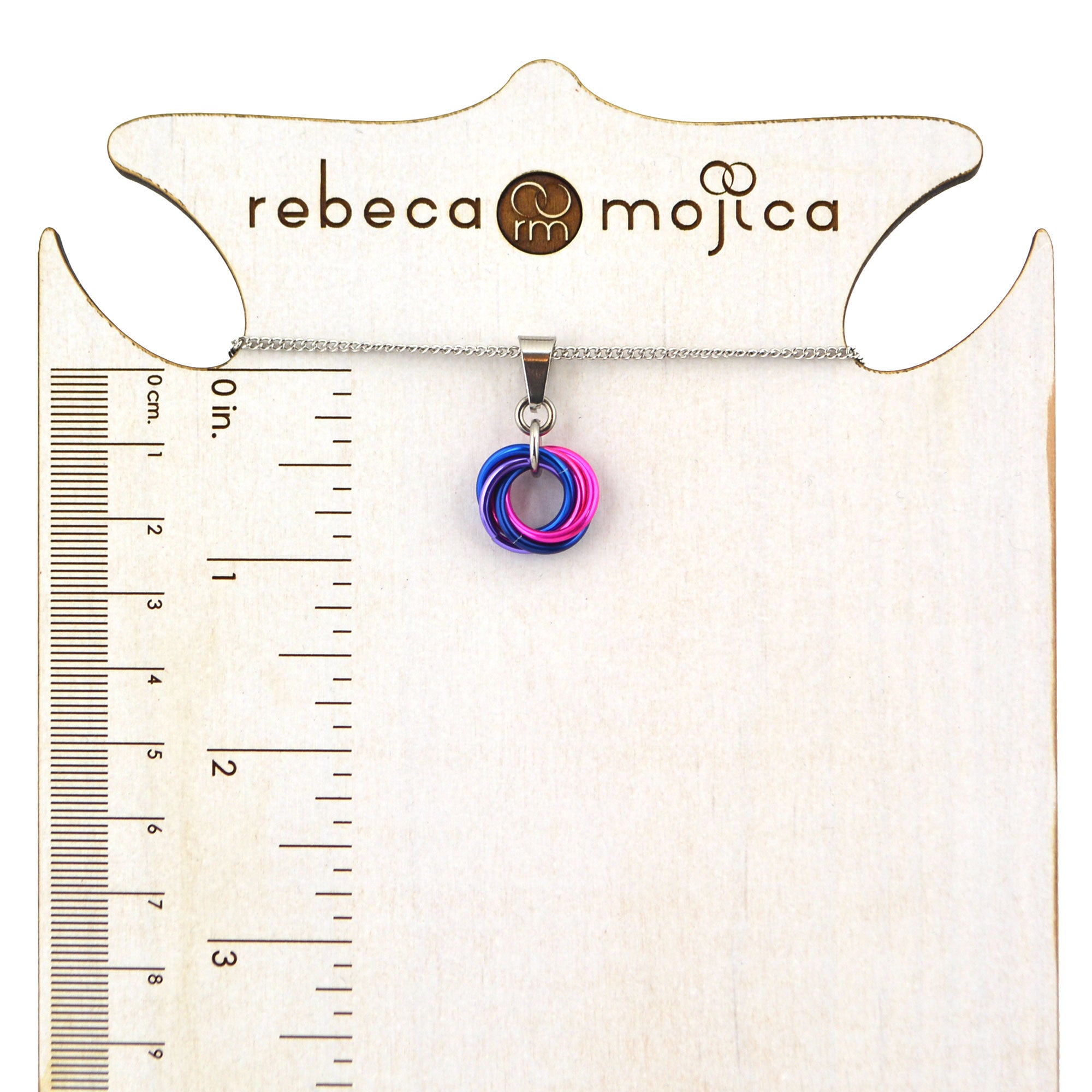 A small bi pride swirl pendant necklace in blue, pink and lilac is displayed on a Rebeca Mojica branded stand with a ruler showing measurements in inches and centimeters. The pendant is approximately 1 inch long