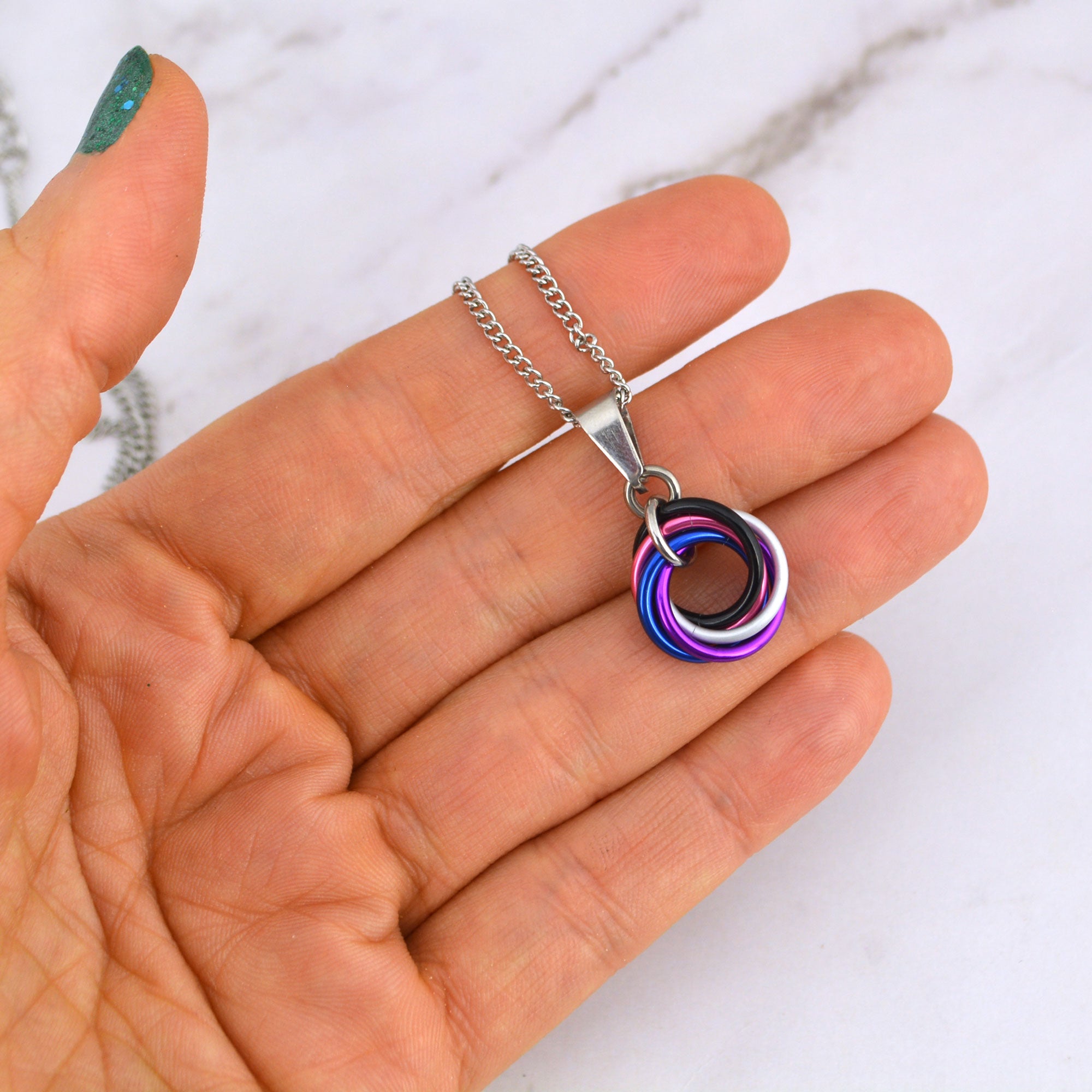 A hand with teal glitter nail polish holds a stainless steel chain necklace featuring a small swirl pendant in the colors of the genderfluid pride flag: black, white, purple, blue and pink. The background is a white marble surface.