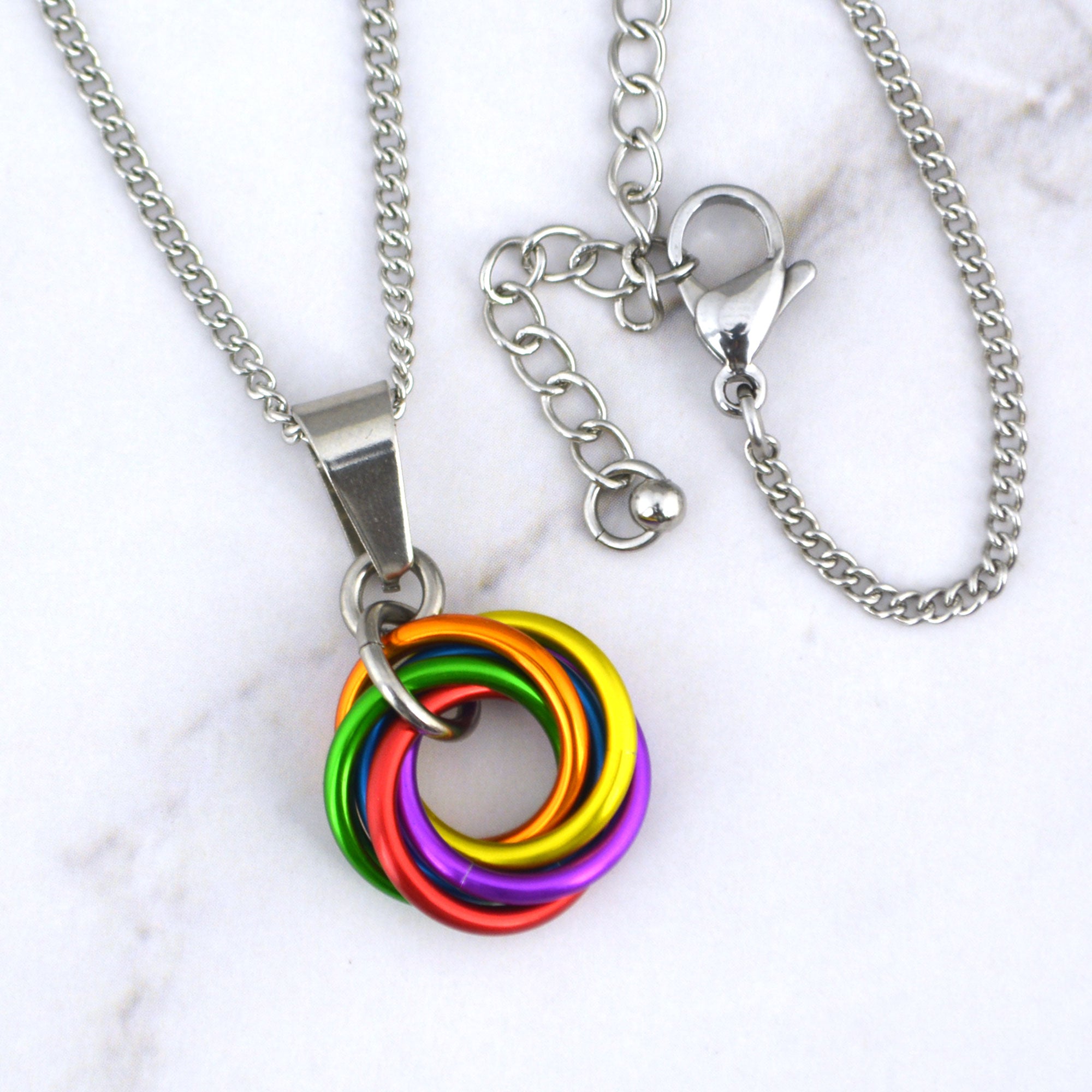 Close-up of a small LGBTQ rainbow pride chainmaille pendant resting on a marble surface. The pendant is a vortex of 6 intertwined rings in red, orange, yellow, green, blue and purple (though the blue ring is mostly hidden under the others).. The links are joined by a steel ring and attached to a smooth steel bail. The lobster claw clasp and extender chain are clearly visible against a white marble background.
