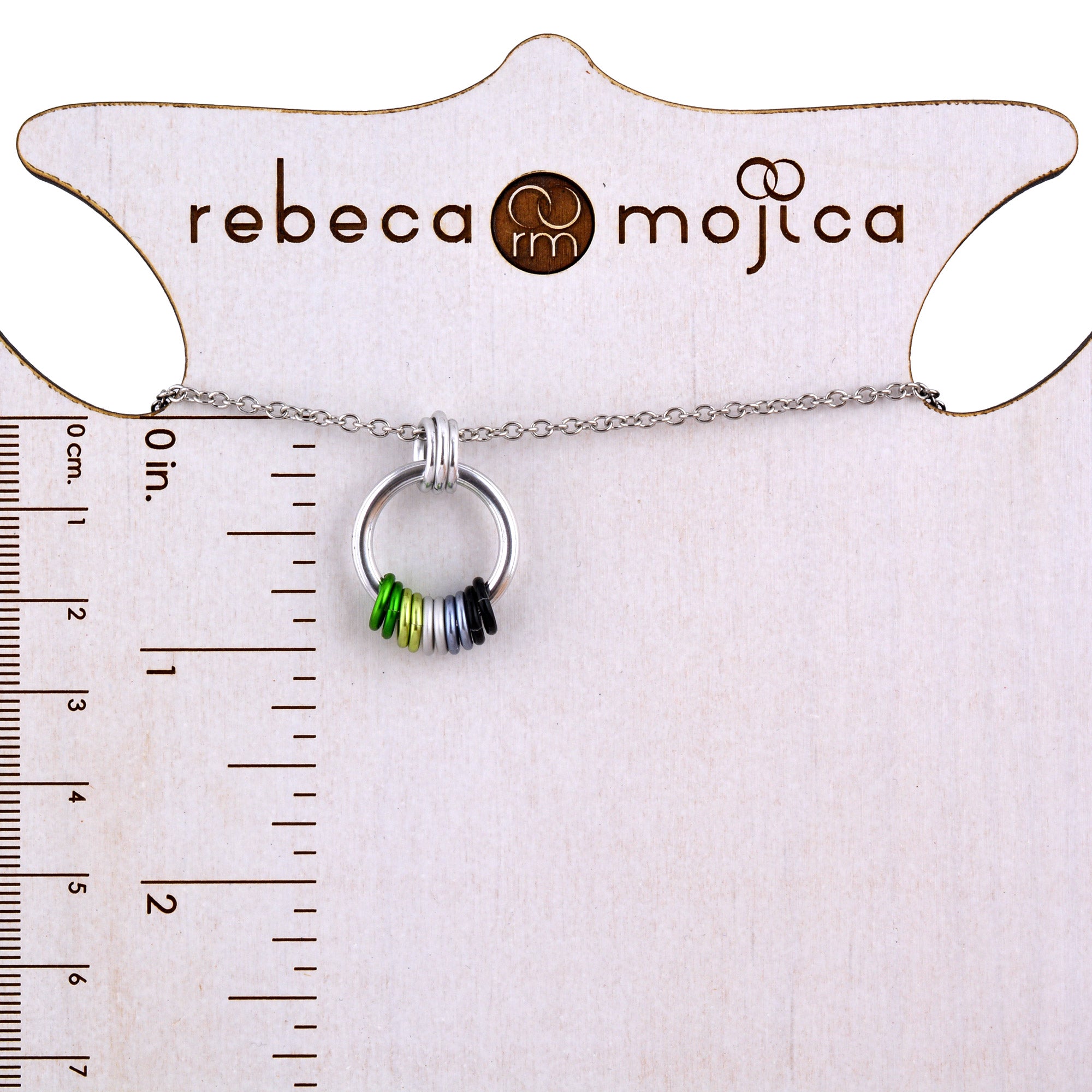 A minimalist Coil Fidget Necklace featuring green, light green, grey, and black rings of the Aromantic Pride flag. The pendant is displayed on a white product card with rulers in inches and centimeters, clearly showing its size.