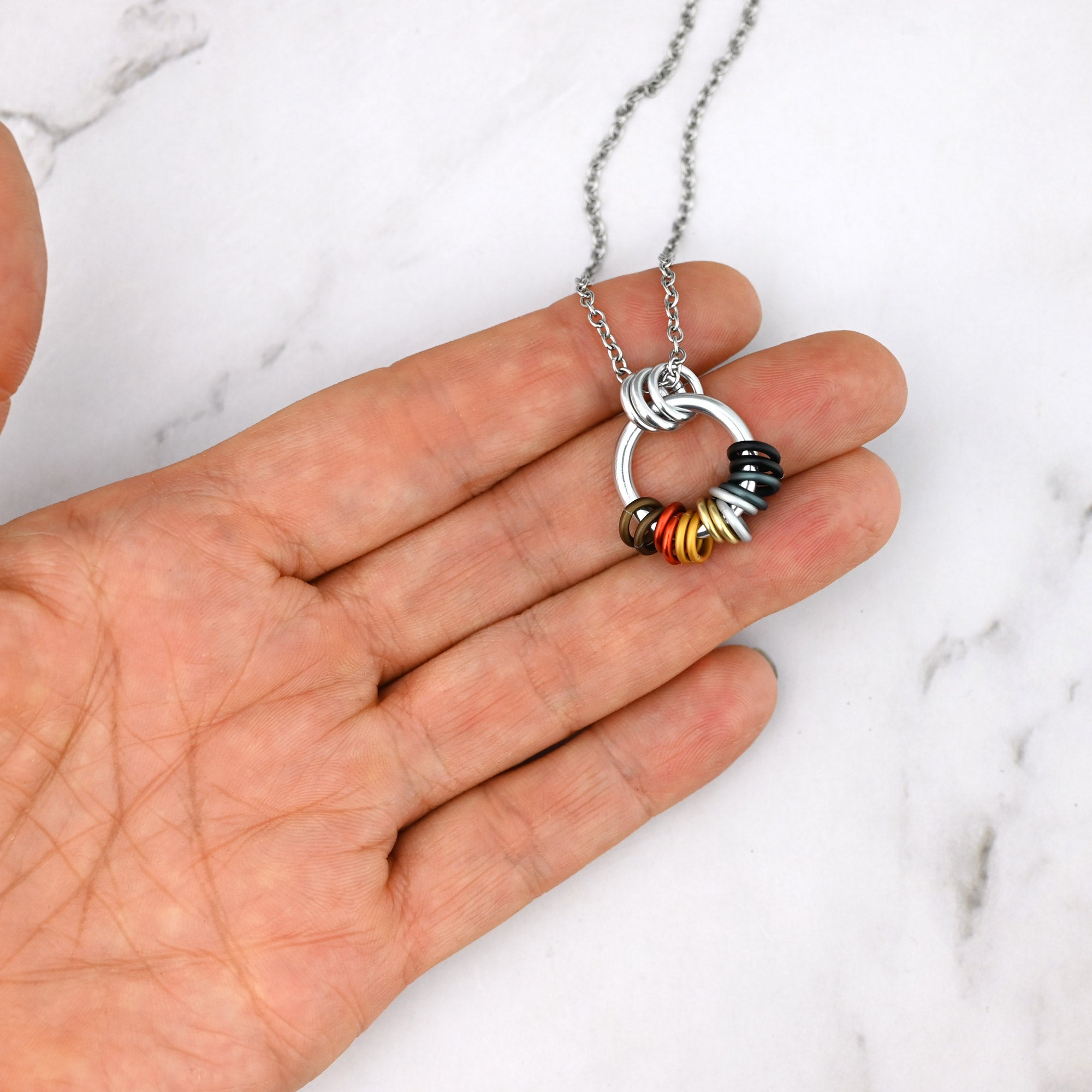 The Bear pride coil necklace held between fingers for scale. The earthy-toned rings sit loosely at the base of the silver hoop, indicating that they can spin and move during wear.