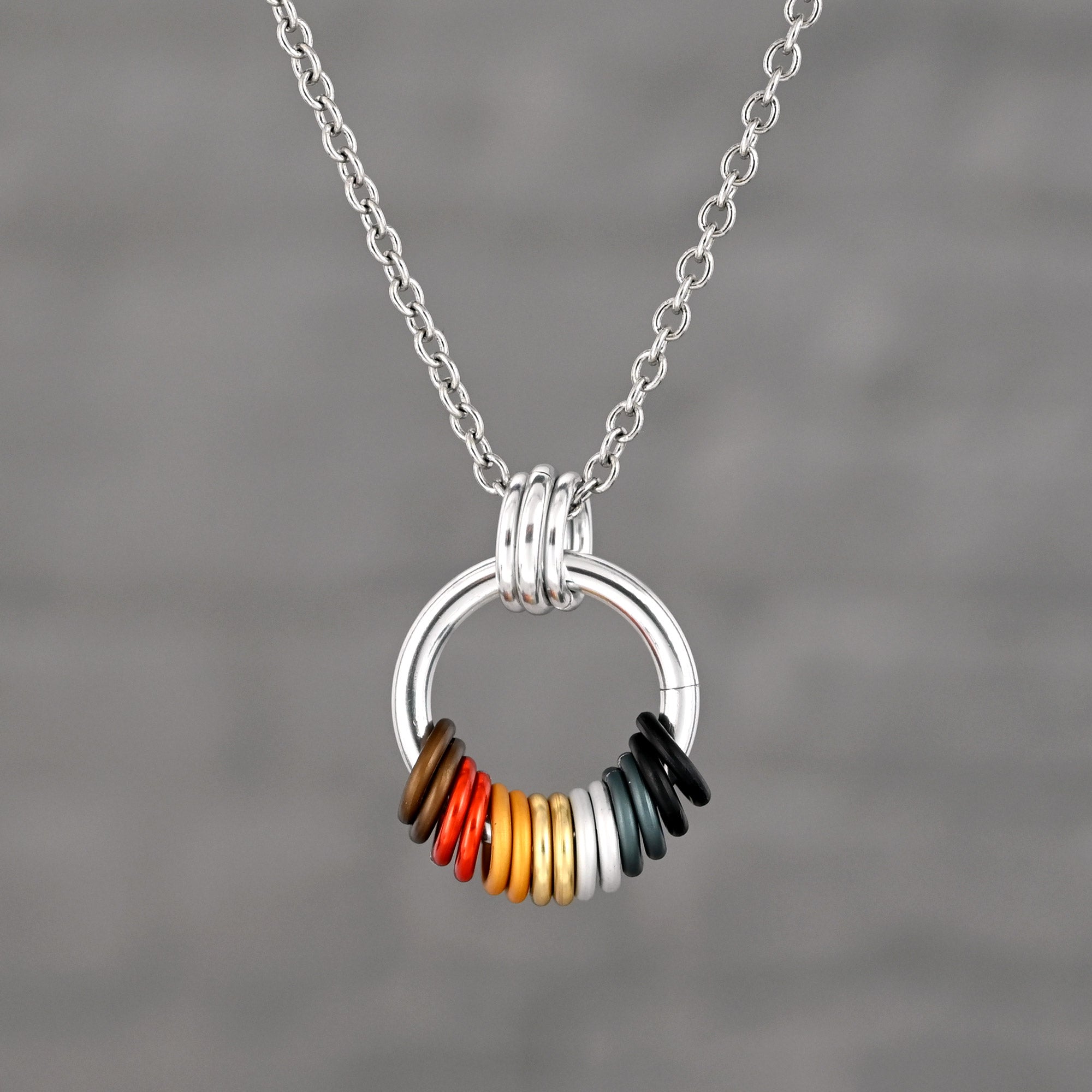 A minimalist silver-tone circular pendant on a stainless steel chain. The base of the hoop features free-spinning aluminum rings in the Bear flag colors: dark brown, rust orange, yellow, tan, white, grey, and black.