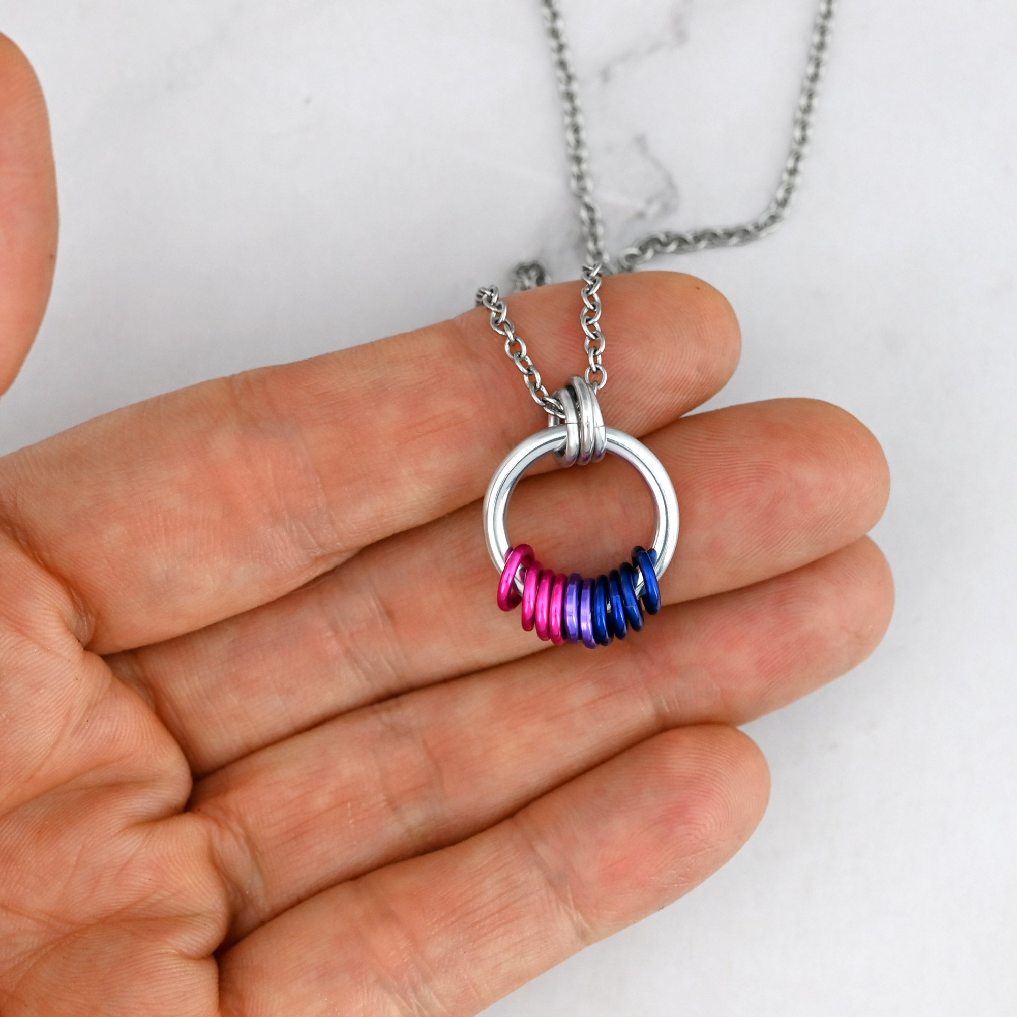A Bi Pride coil pendant is held against fingertips to show its compact size. The pink, purple, and blue rings are grouped at the bottom of the hoop, with visible space indicating they are free-spinning and tactile.