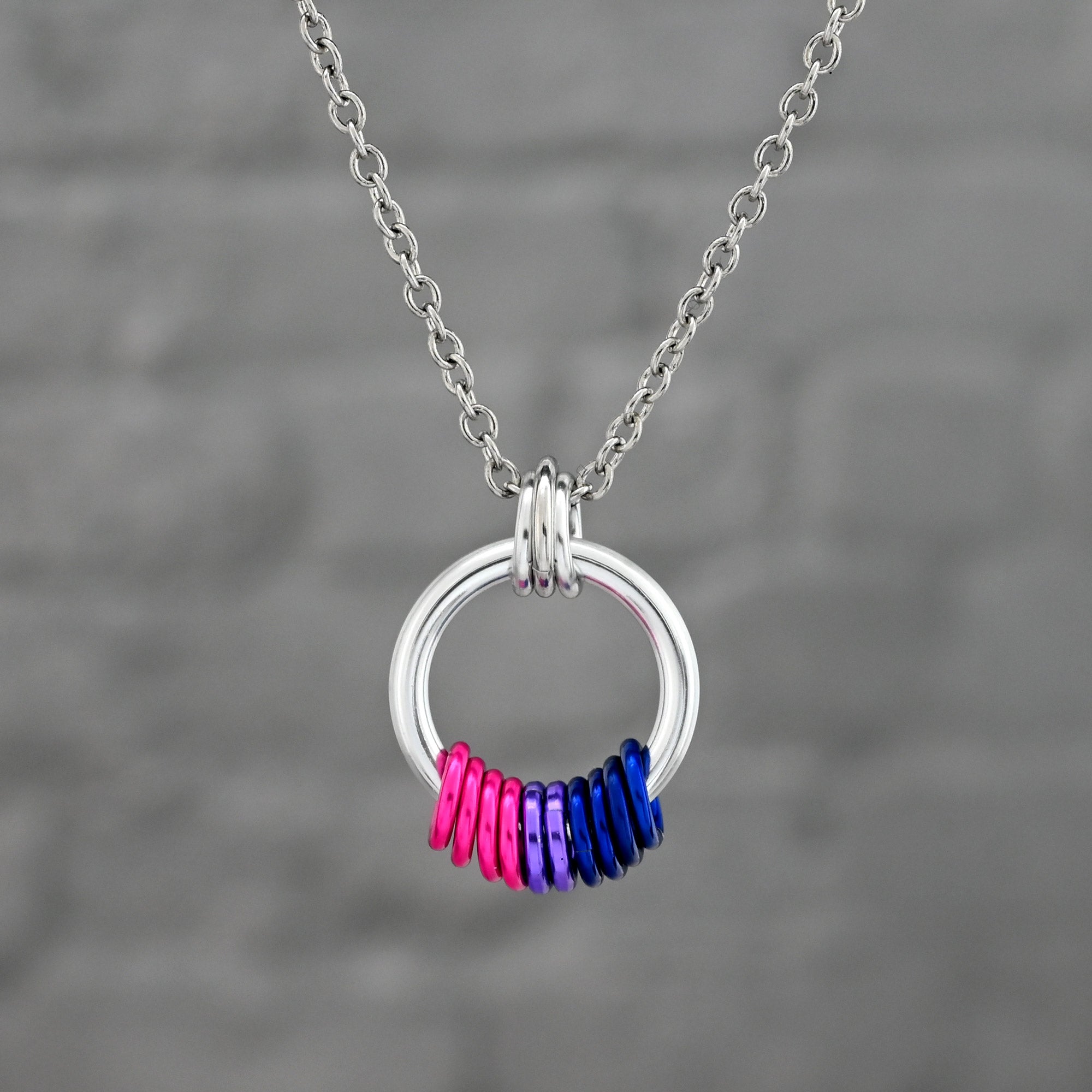 A silver-tone loop pendant with a coil of free-spinning rings in Bisexual Pride colors: deep pink, purple, and royal blue. The pendant hangs from a sturdy stainless steel link chain.