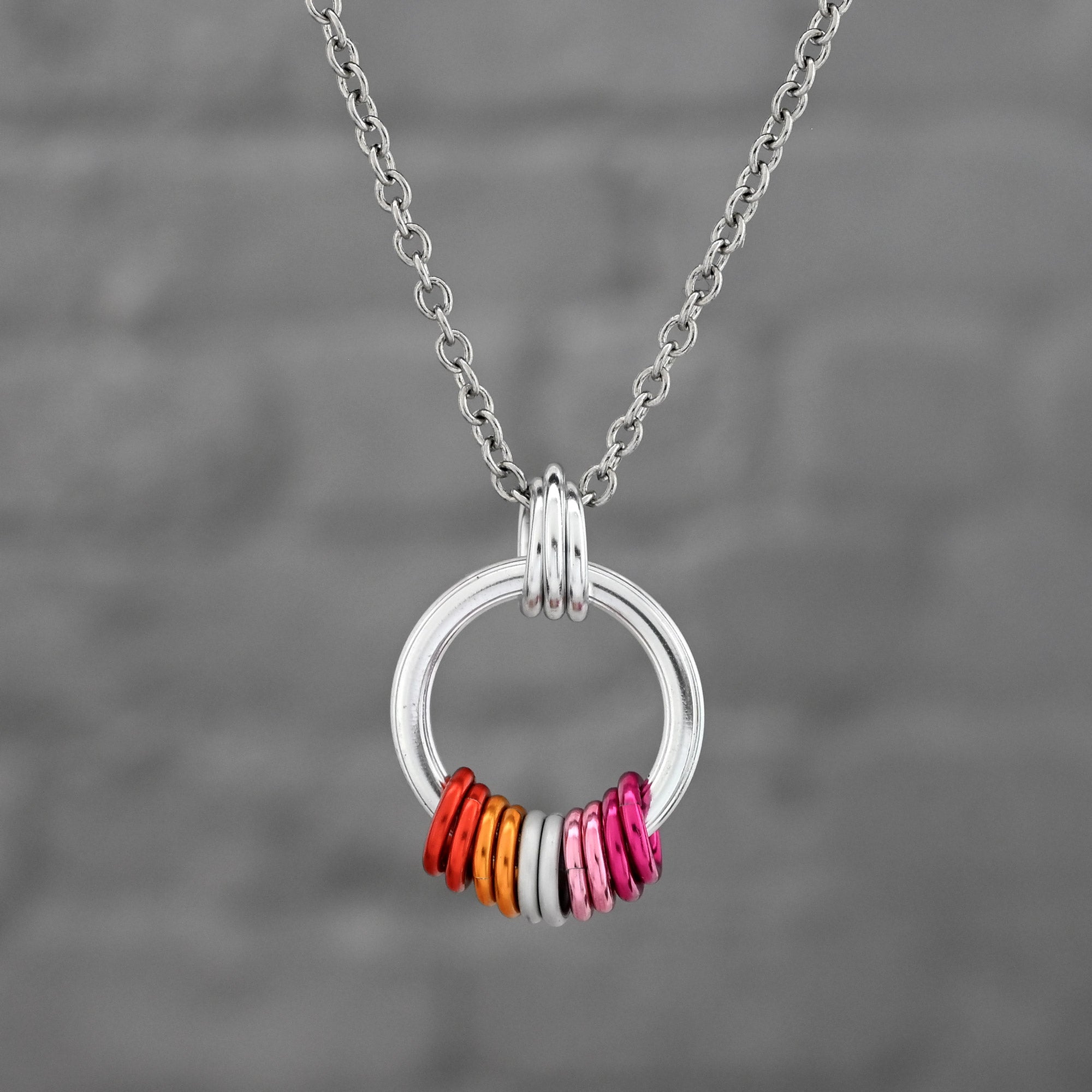 A minimalist silver pendant featuring a series of colorful rings inspired by the Lesbian Pride flag. The rings transition through shades of dark orange, light orange, white, light pink, and dark pink.
