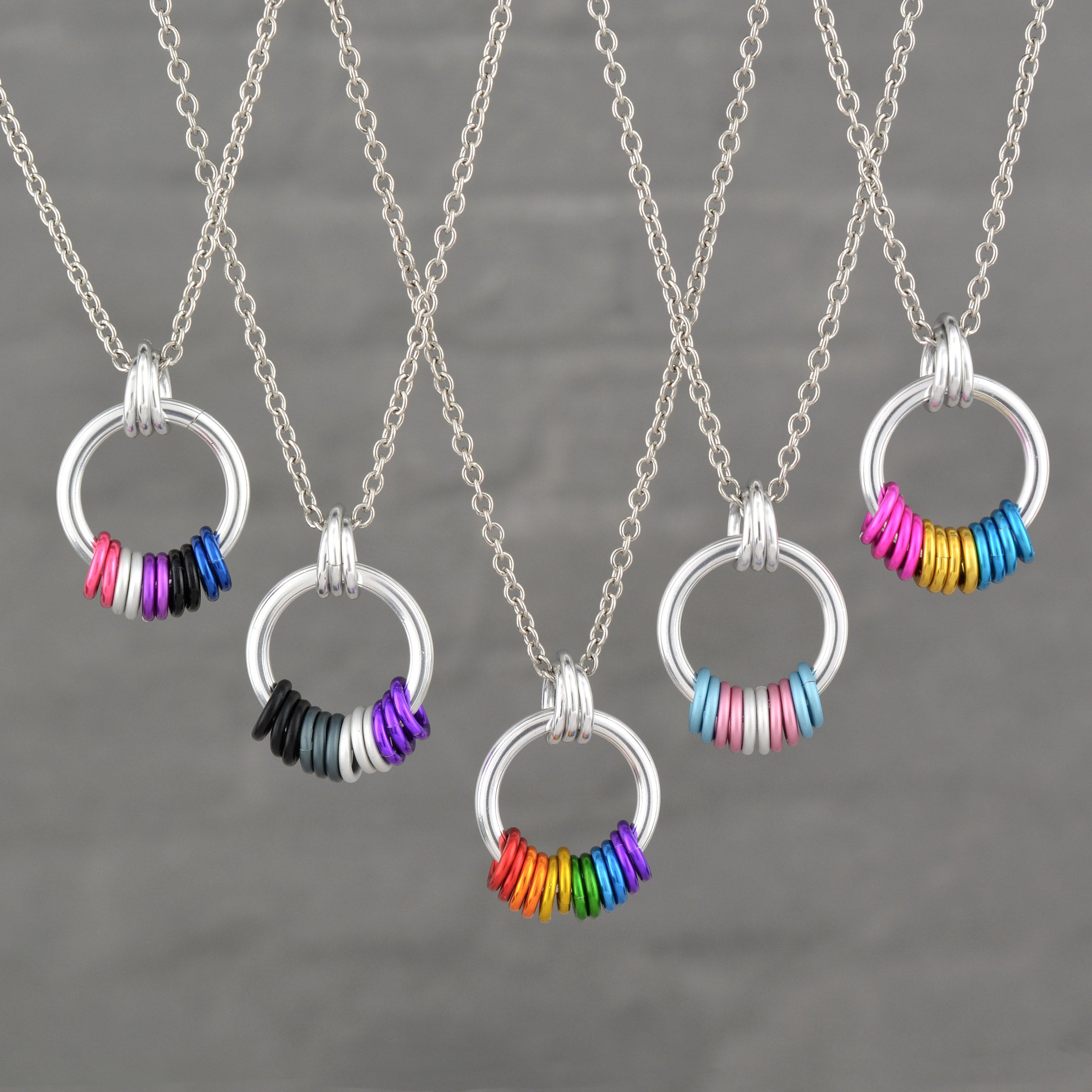 A group of five distinct Pride Coil Fidget Necklaces, each showcasing different flag colors (from left: Genderfluid, Asexual, Rainbow, Transgender, Pansexual). The silver coil pendants with their freely spinning aluminum rings are displayed against a neutral grey background.
