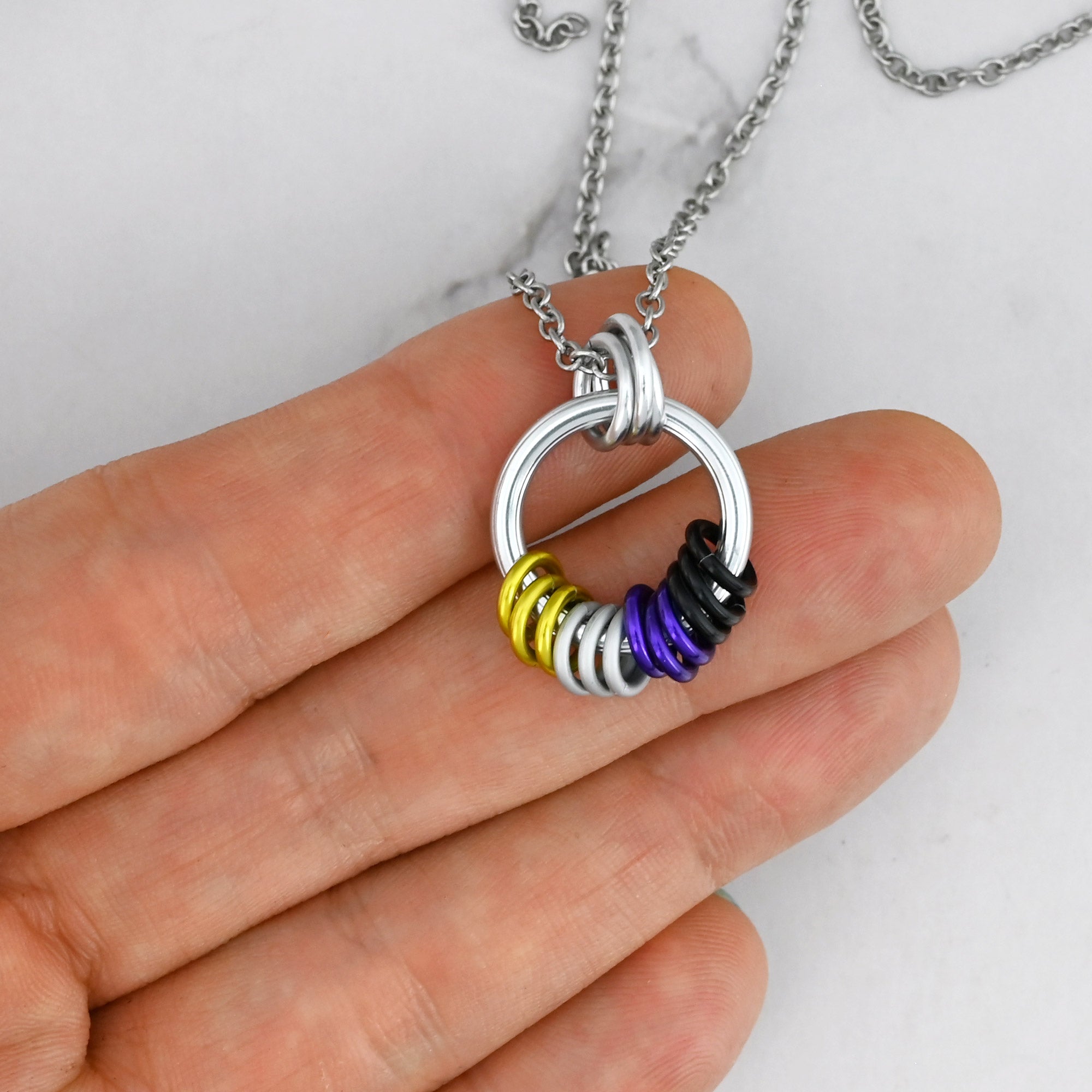 The Nonbinary Pride necklace held against a person's fingertips, illustrating minimalist aesthetic of the jewelry. The small yellow, white, purple and black rings re clustered at the bottom of the silver ring, and sitting in such a way to indicate the tactile movement of the chainmaille links.