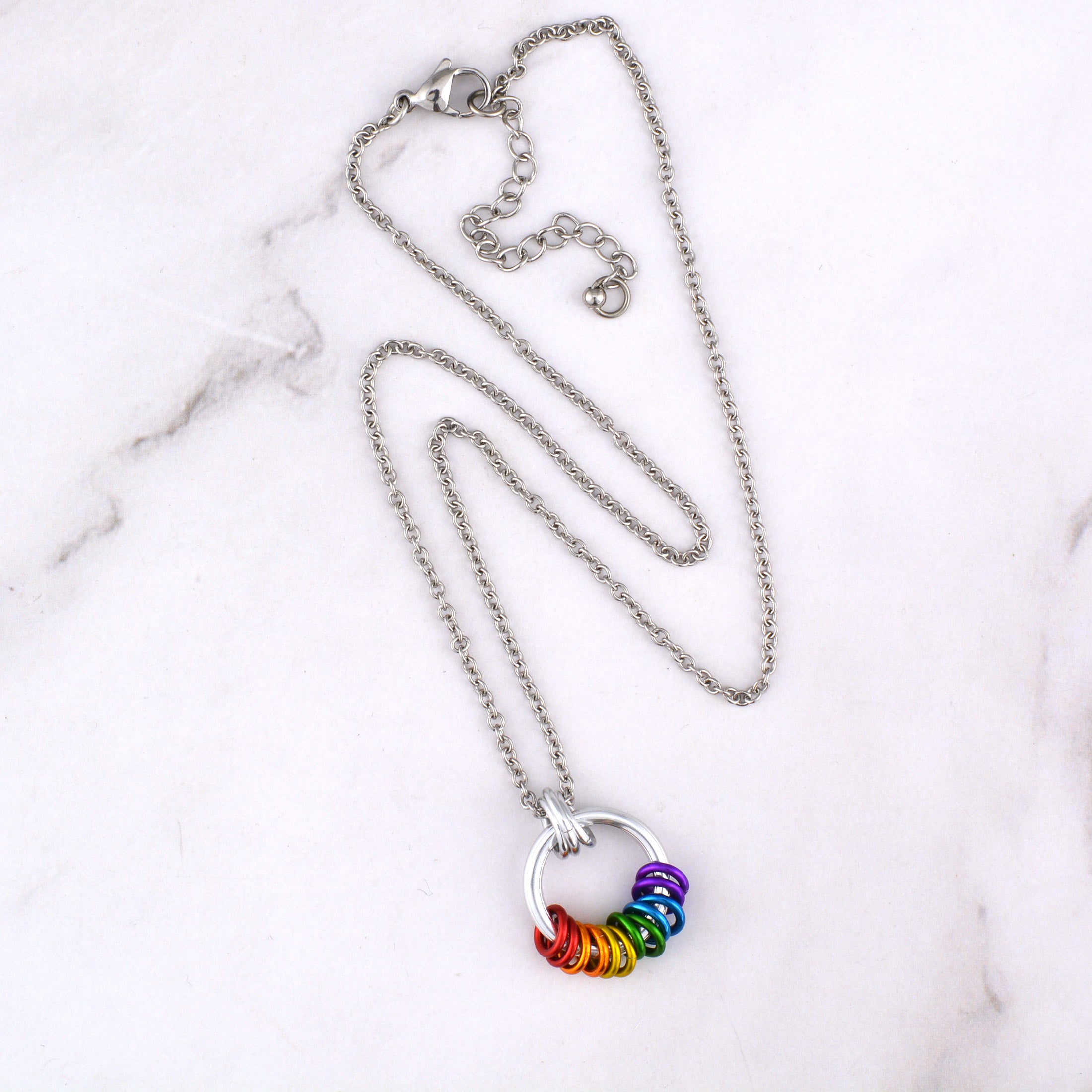An overhead shot of the Rainbow Pride Coil Fidget Necklace with its stainless steel chain laid out on a white marble surface. The image clearly displays the silver coil pendant, vibrant rainbow rings, and the lobster claw clasp.