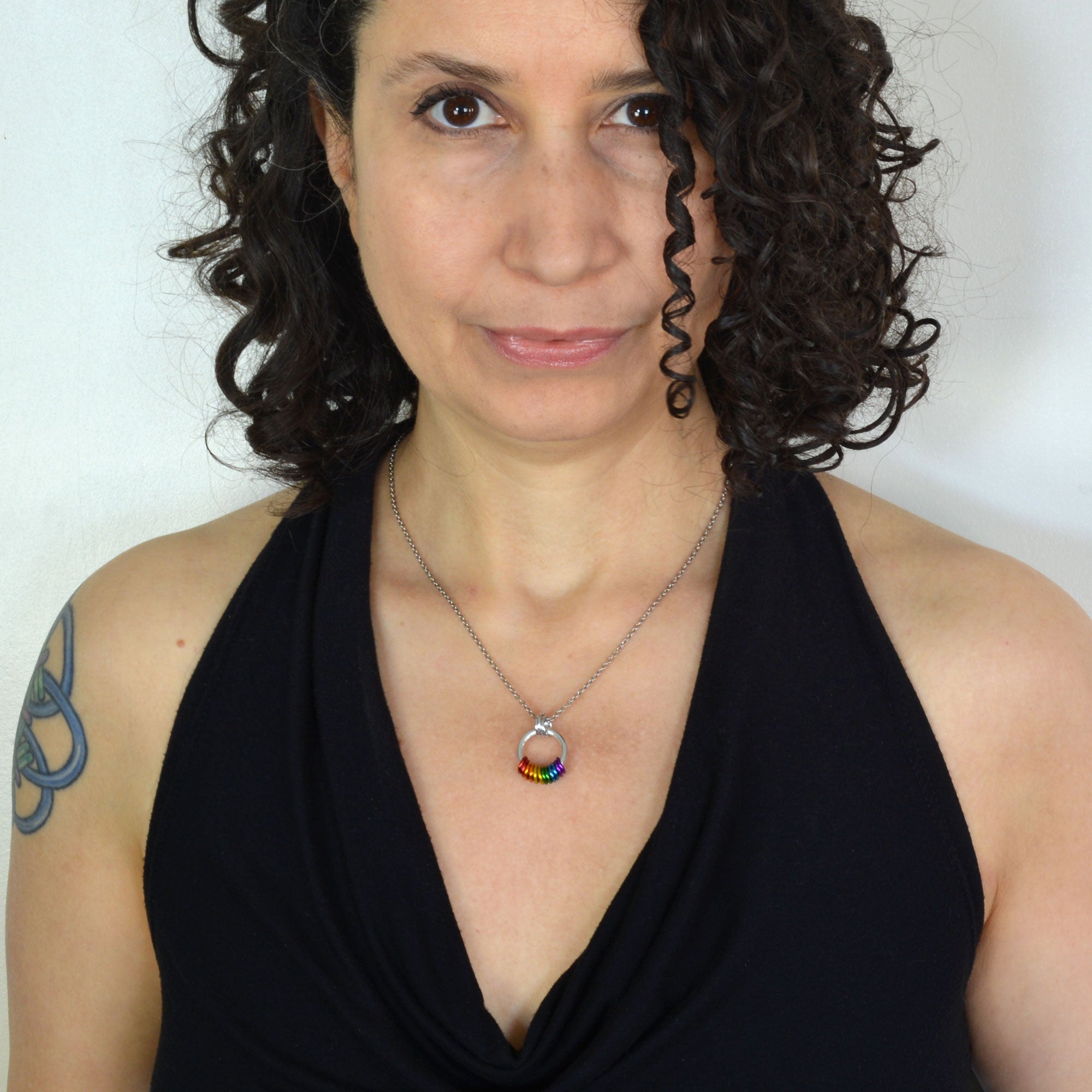 Rebeca Mojica, a chainmaille artisan, modeling the Rainbow Pride Coil Fidget Necklace. She wears a black top and has a subtle tattoo on her arm. Her shoulder-length curly dark brown hair frames her face as she looks directly at the camera with a slight smile. The necklace features bright rainbow-colored rings, worn discreetly.