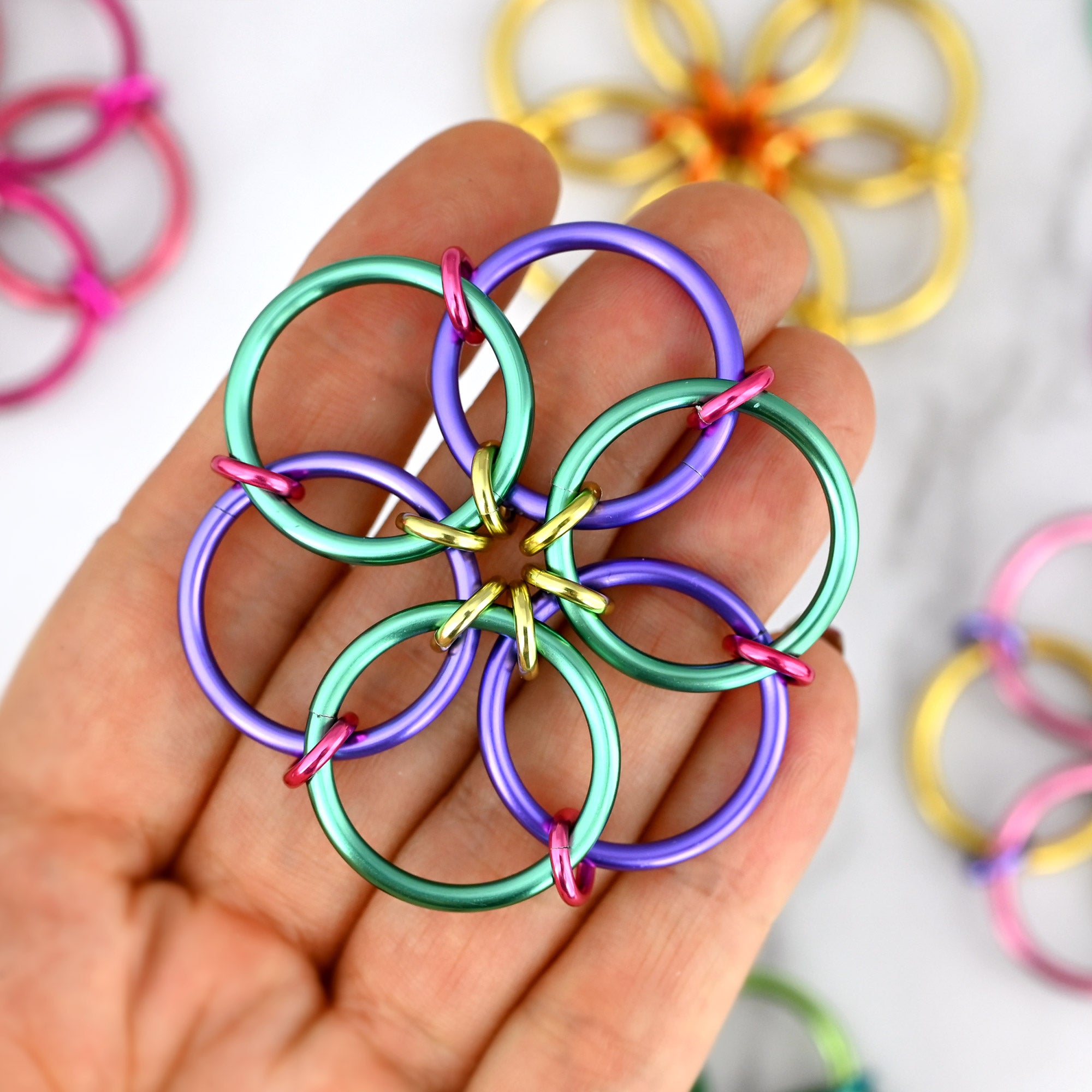 A close-up photograph of the "Cool Blossom" Fidget Flower being held on a person’s fingertips to show its size. This artisan chainmaille fidget features large purple and light teal outer rings, joined by pink and light gold small connector rings. In the soft-focus background, the other colorways from the "Vol 1: Fresh Bloom" collection are visible on a white marble surface, highlighting the bright spring colors and handcrafted mandala shape of the sensory accessory.