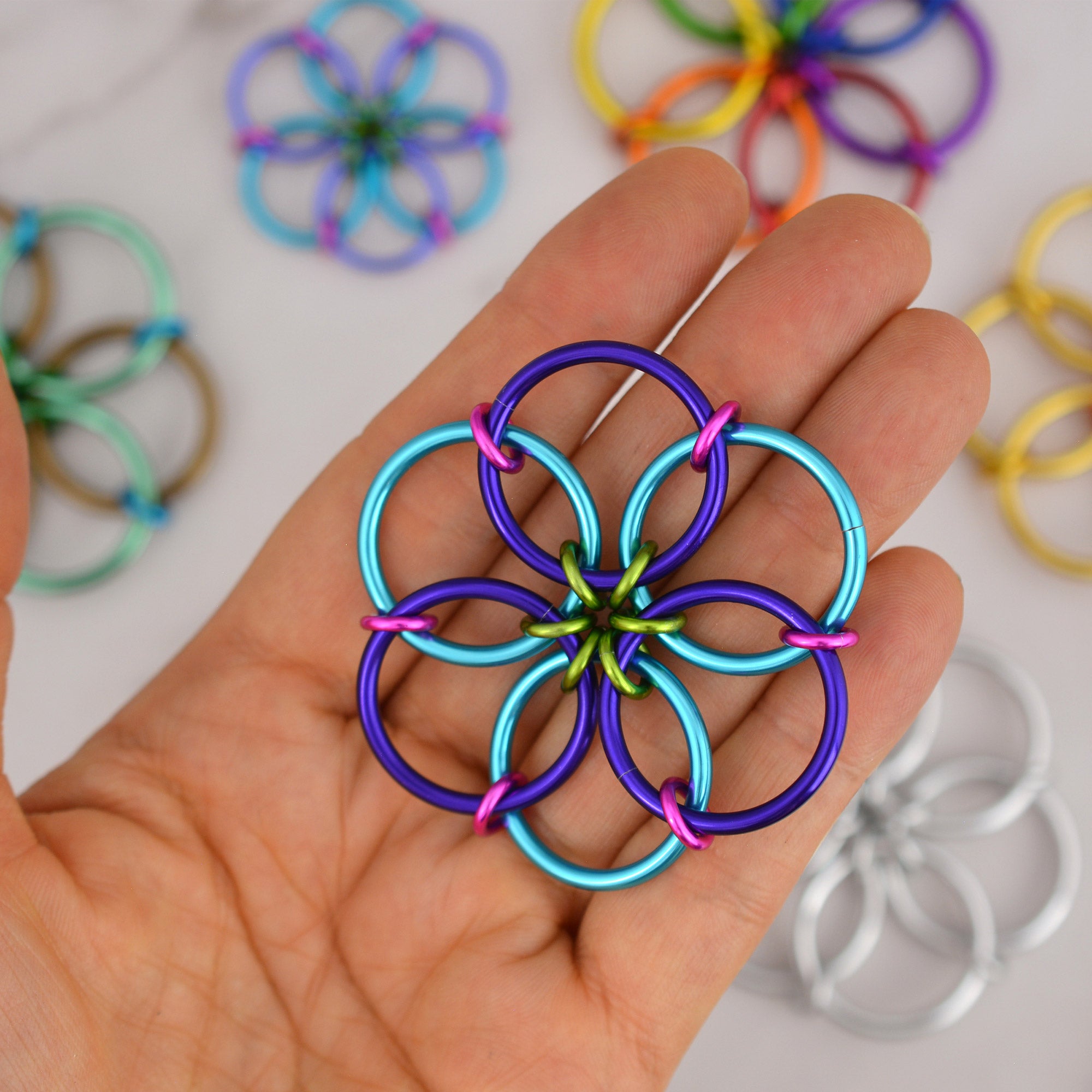 The Funfetti Fidget Flower, with rings in turquoise, purple, lime green, and hot pink, held flat in the palm of a hand. Other Fidget Flowers in various colors are blurred in the background.