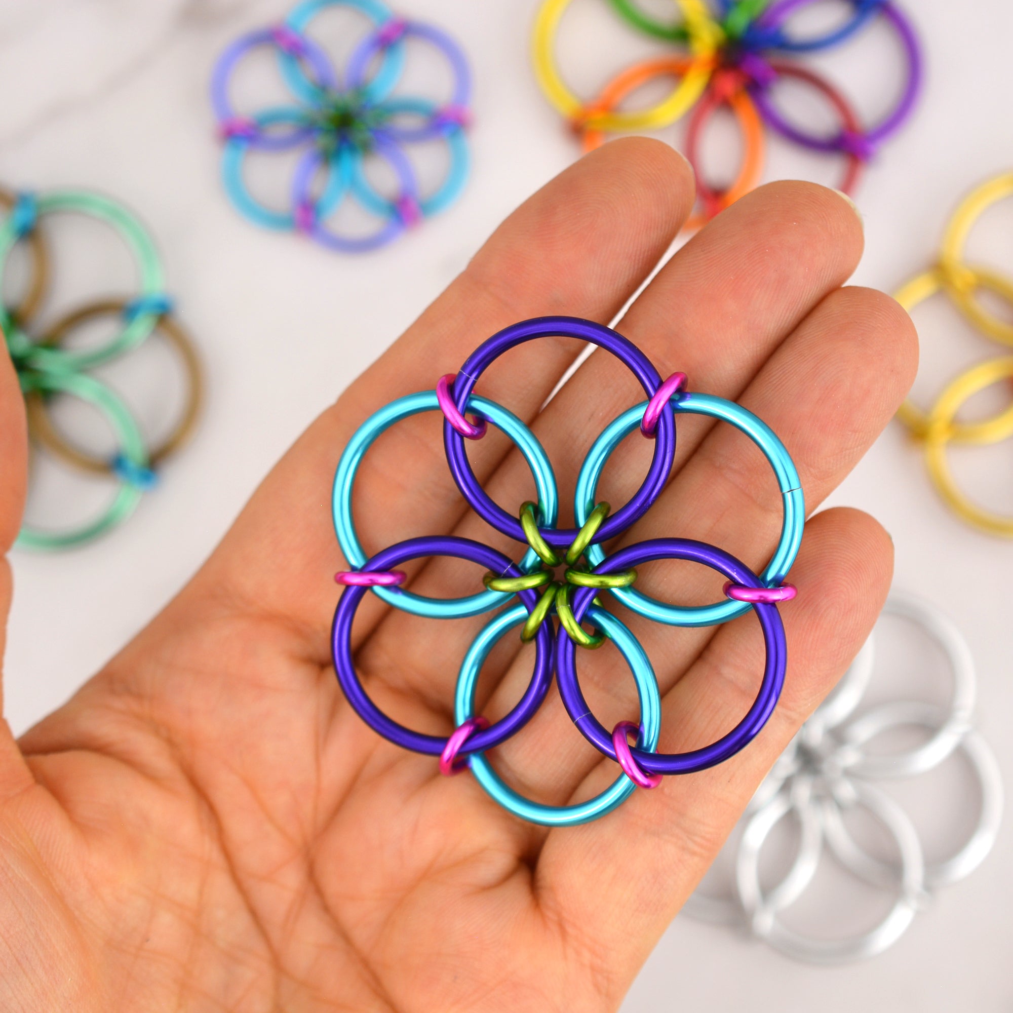 The Funfetti Fidget Flower, with rings in turquoise, purple, lime green, and hot pink, held flat in the palm of a hand. Other Fidget Flowers in various colors are blurred in the background.