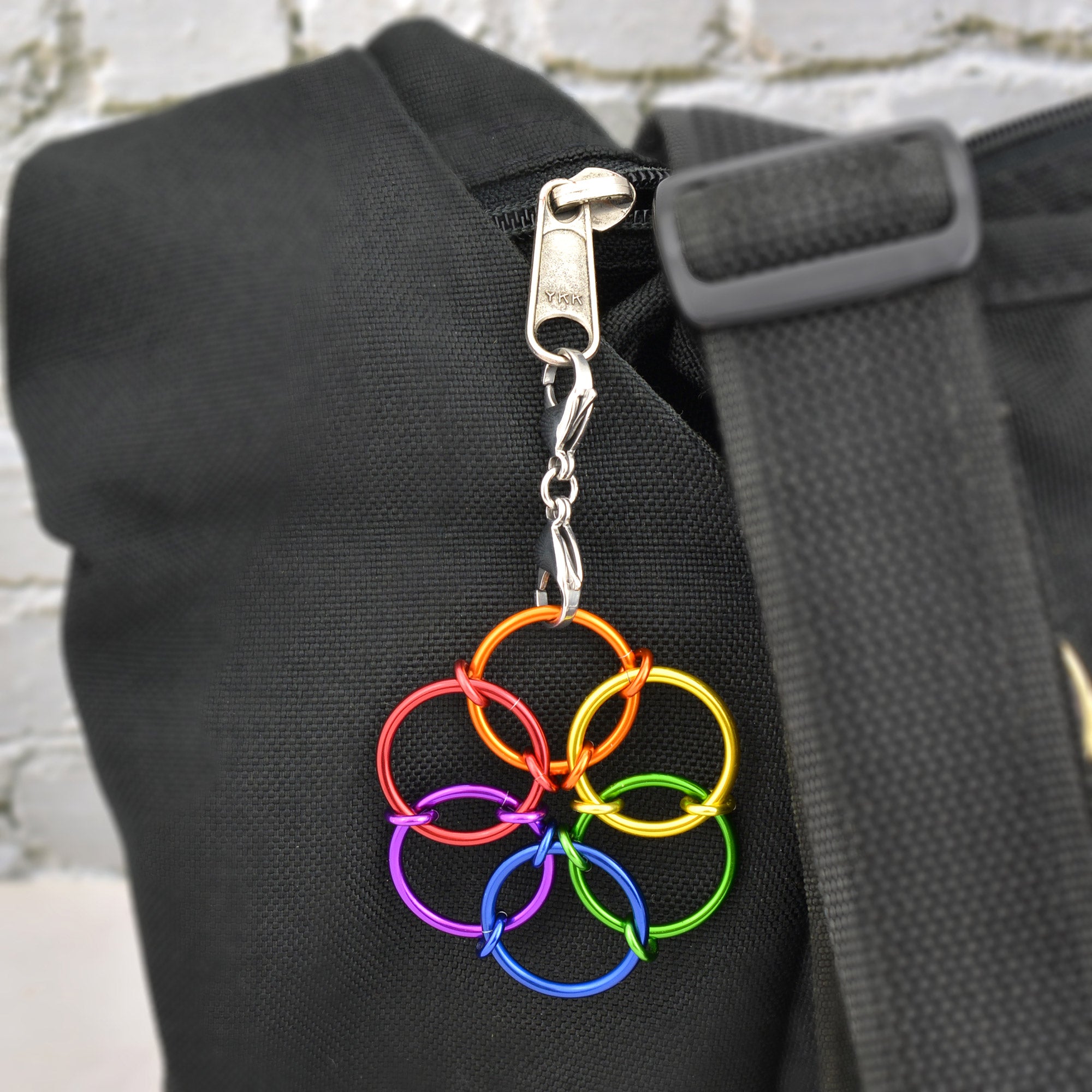 A rainbow-colored Fidget Flower charm attached to the zipper of a black backpack or bag strap using a small silver clip. The flower has rings in red, orange, yellow, green, blue, and purple.