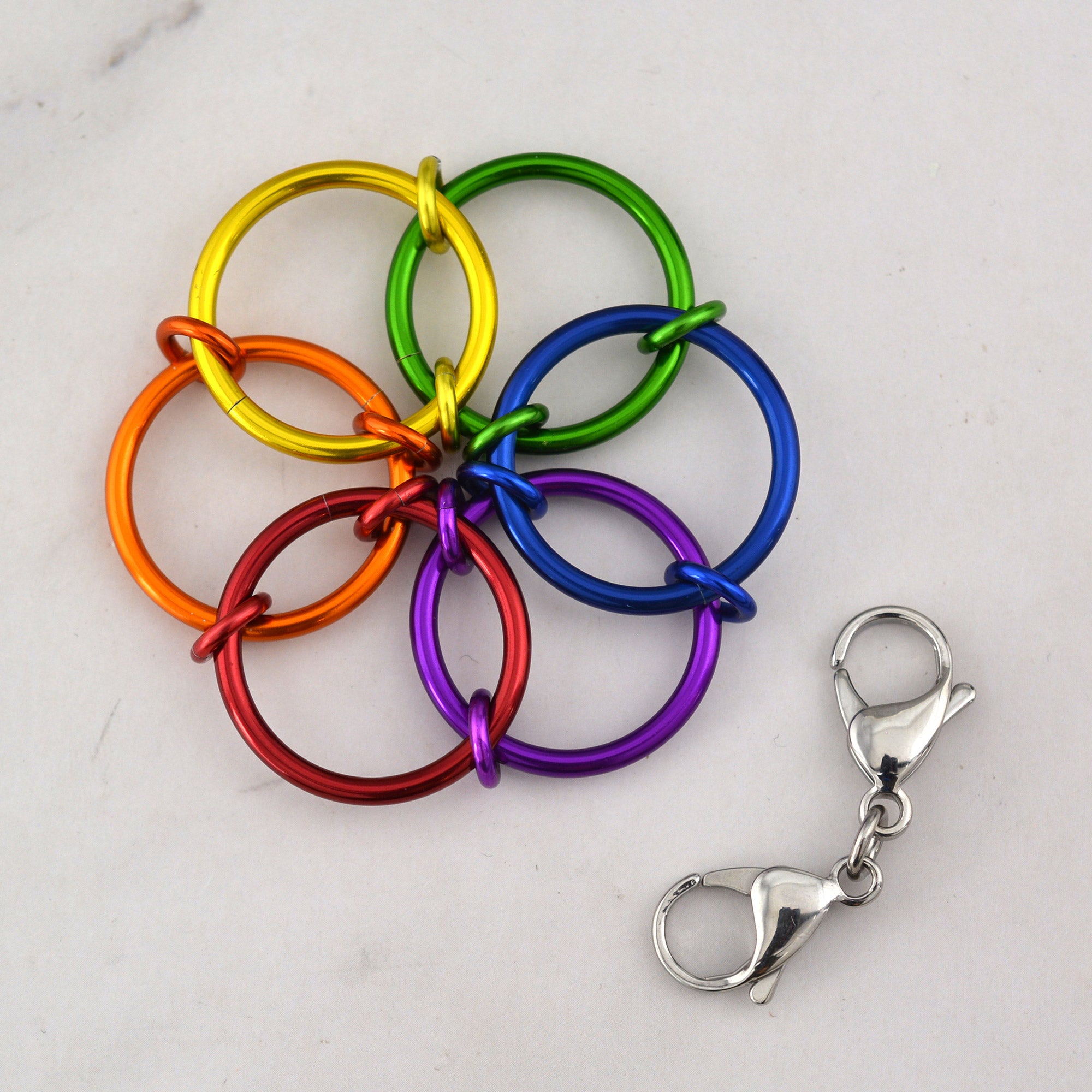 A close-up aerial view of the Funfetti Fidget Flower pendant, featuring interlocking rings in vibrant turquoise and deep purple, accented by smaller lime green and hot pink rings in the center, with a steel chain attached.