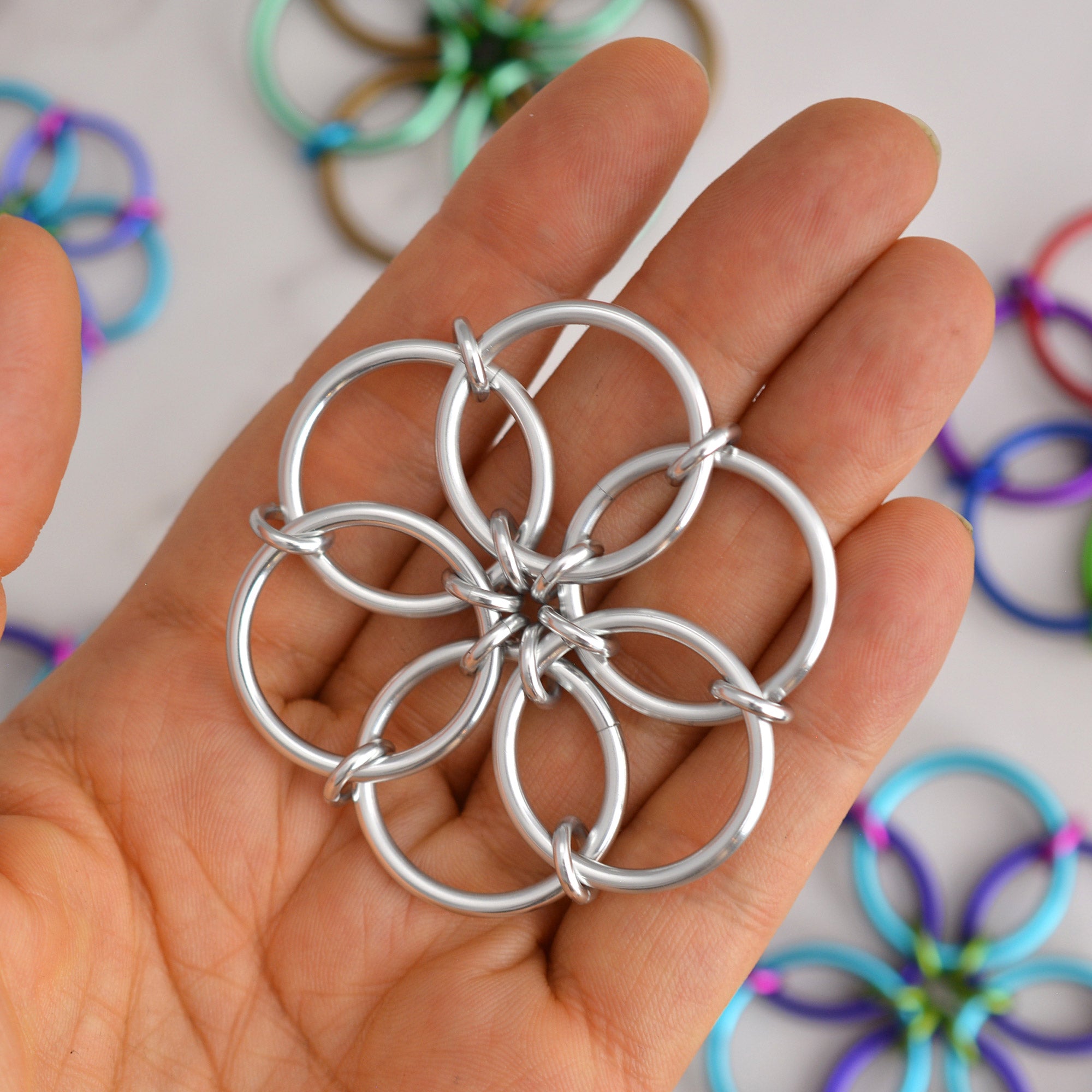 silver Fidget Flower held flat in the palm of a hand, showing the size and detail of the interlocking aluminum rings. Other colored Fidget Flowers are blurred in the background.