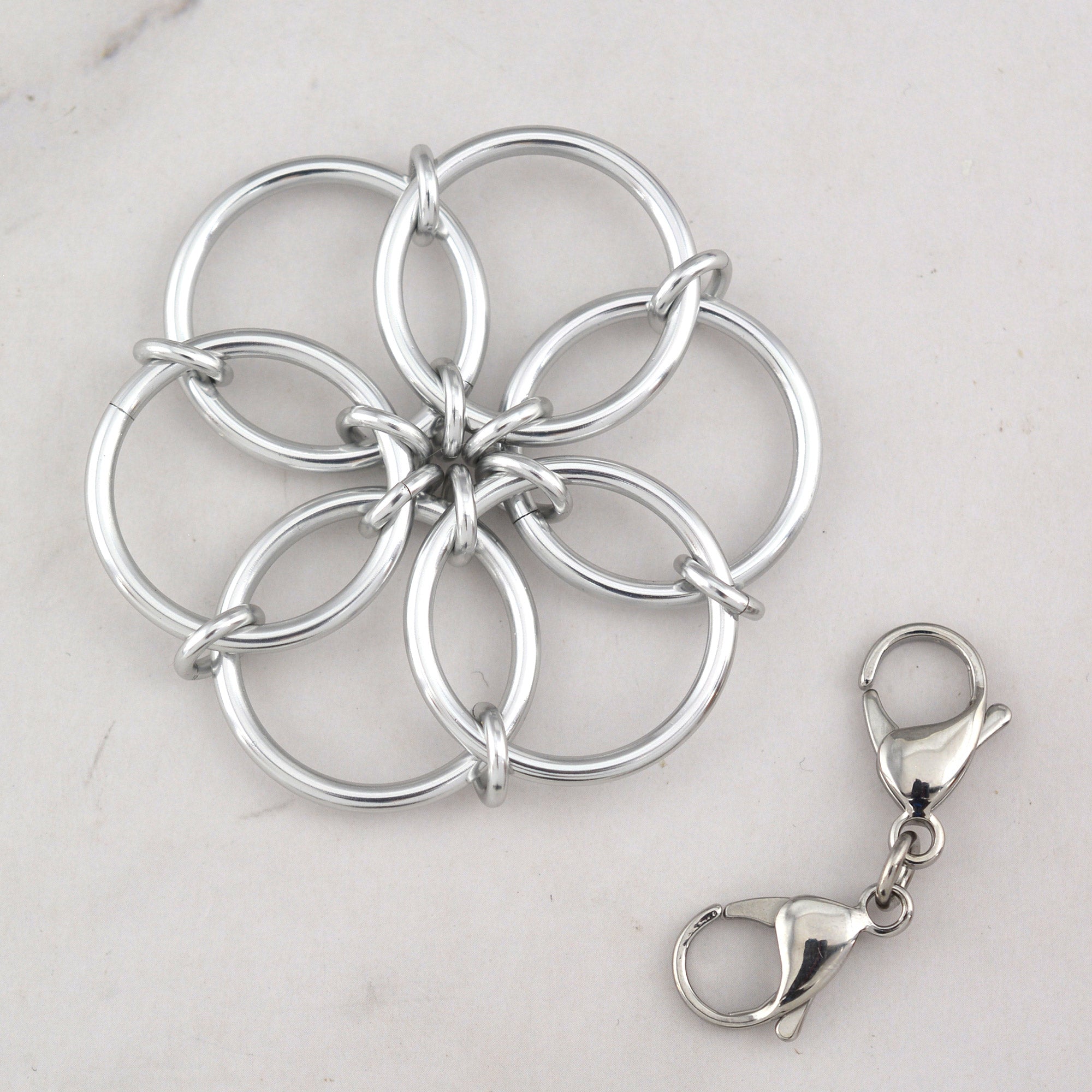 The silver Fidget Flower accessory lying flat next to a detached double-ended silver lobster claw clip, which is used to attach the flower to bags or zippers.