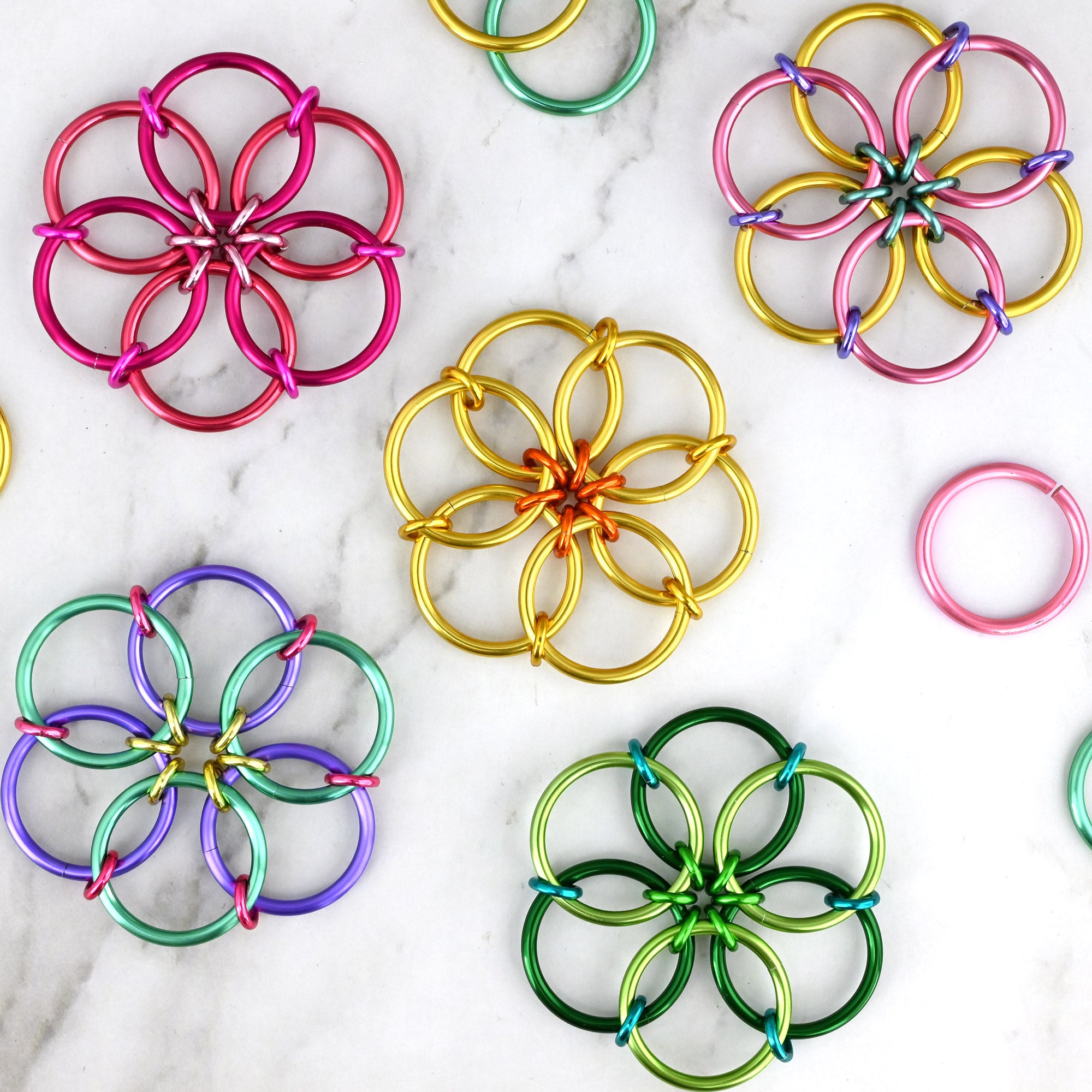 A top-down product photograph showing 5 colorful flower-shaped chainmaille fidgets arranged on a white marble background. Each flexible, circular 'mini mandala' features different spring-themed colorways in shades of pink, green, lilac and gold.