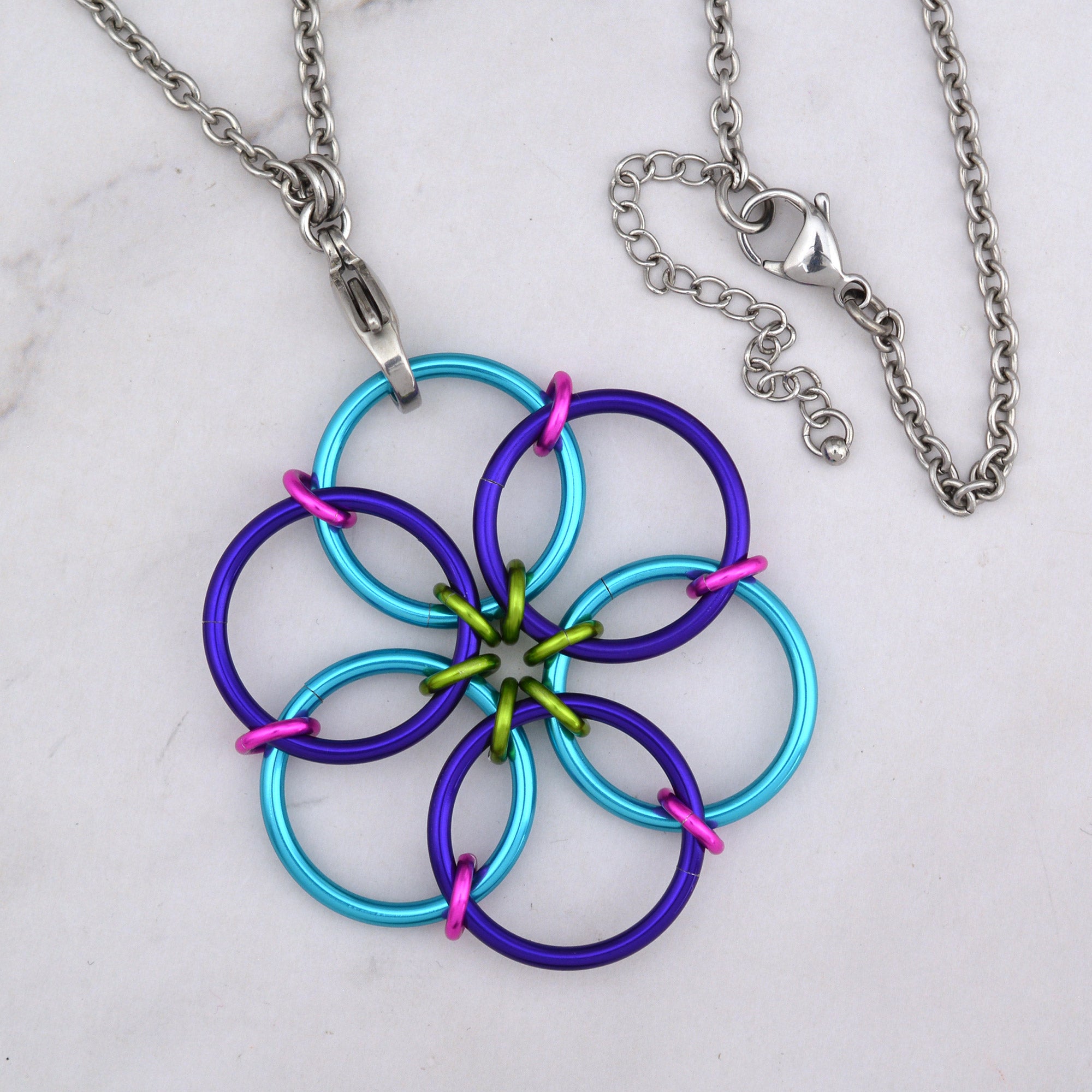 A close-up aerial view of the Funfetti Fidget Flower pendant, featuring interlocking rings in vibrant turquoise and deep purple, accented by smaller lime green and hot pink rings in the center, with a steel chain attached.