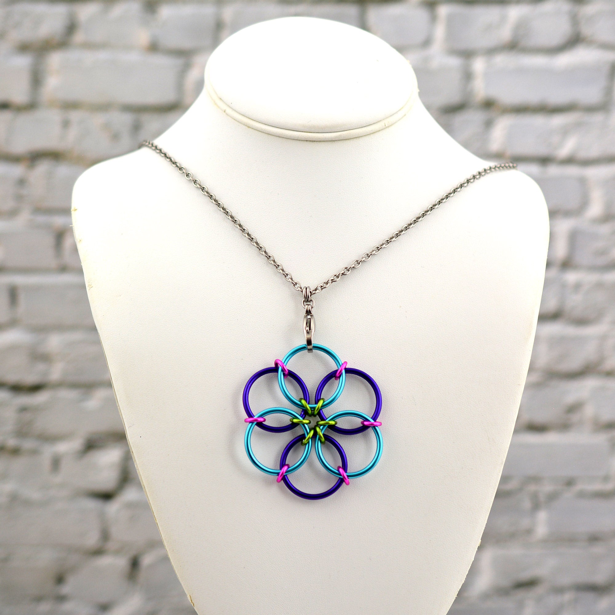 The Funfetti Fidget Flower necklace, featuring turquoise, purple, lime green, and hot pink rings, displayed on a white jewelry neck form against a white brick background.