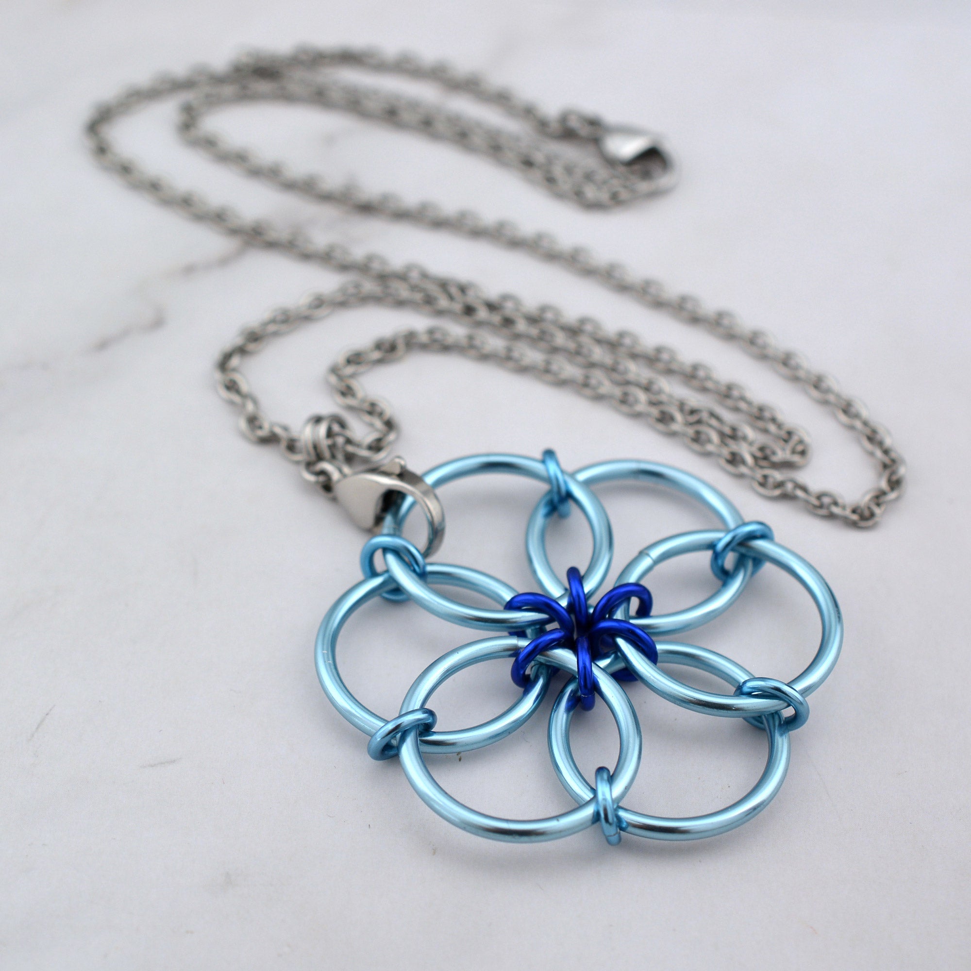 An angled aerial view of the Light Blue Fidget Flower necklace lying on a white marble surface, showing the subtle blend of light blue rings with deep royal blue accent rings at the center.