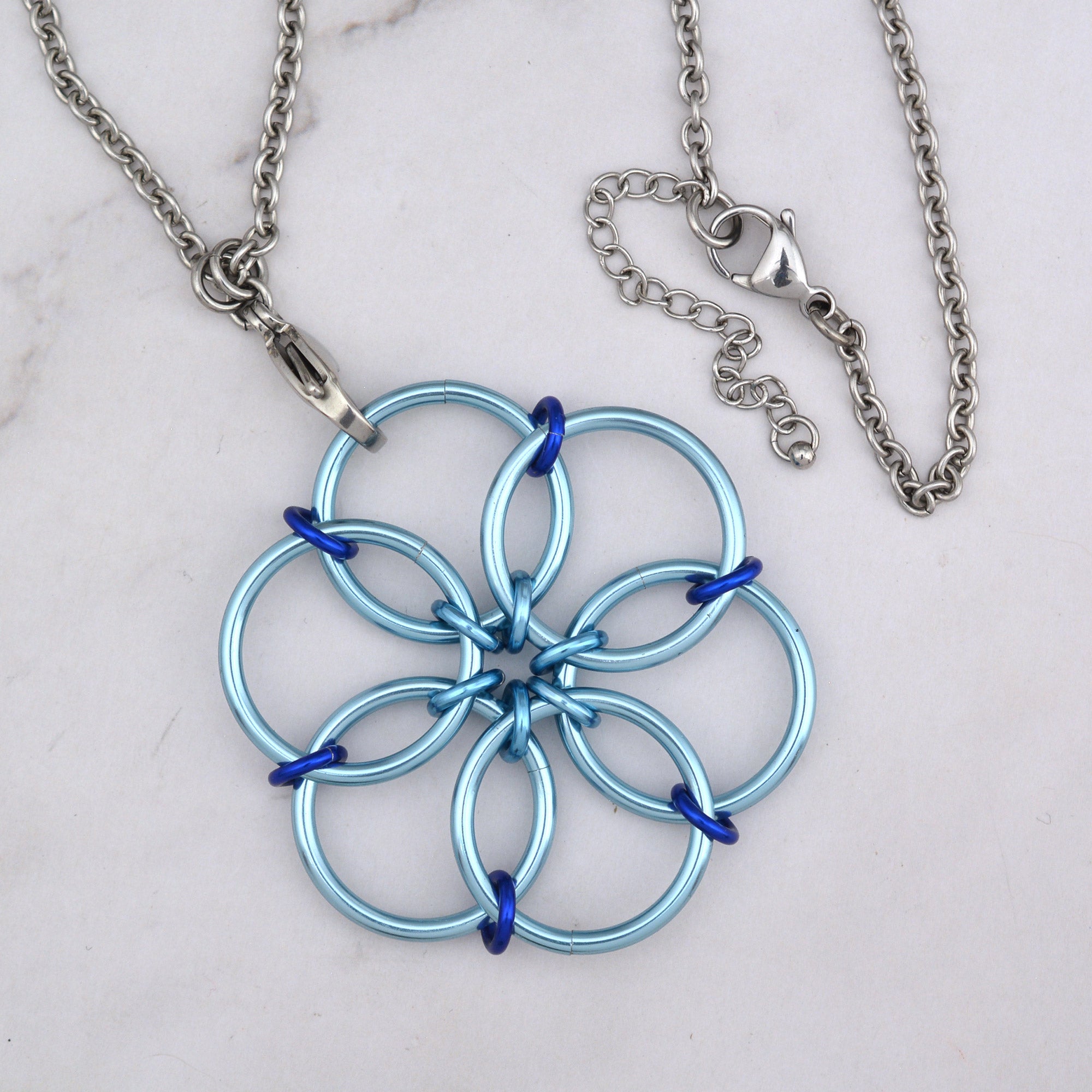 A close-up aerial view of the Light Blue Fidget Flower pendant, highlighting the interlocking light blue rings and the small, dark royal blue accent rings connecting them. The stainless steel chain is visible to the side.