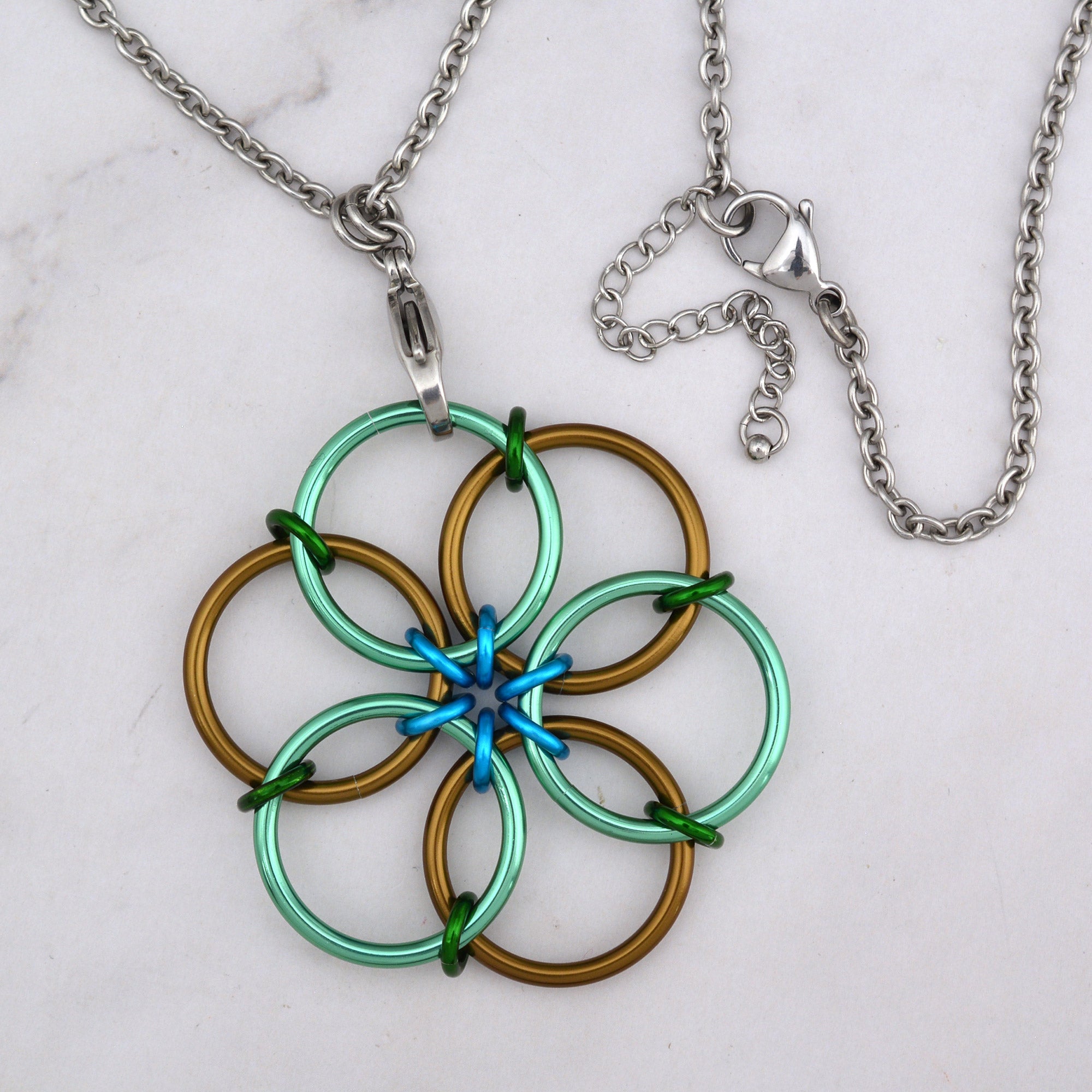 A close-up aerial view of the Magical Woodland Fidget Flower pendant, showcasing the large mint green and russet brown rings, with small forest green and turquoise rings in the center connecting them. The stainless steel lobster claw clasp and extender is visible at upper right.