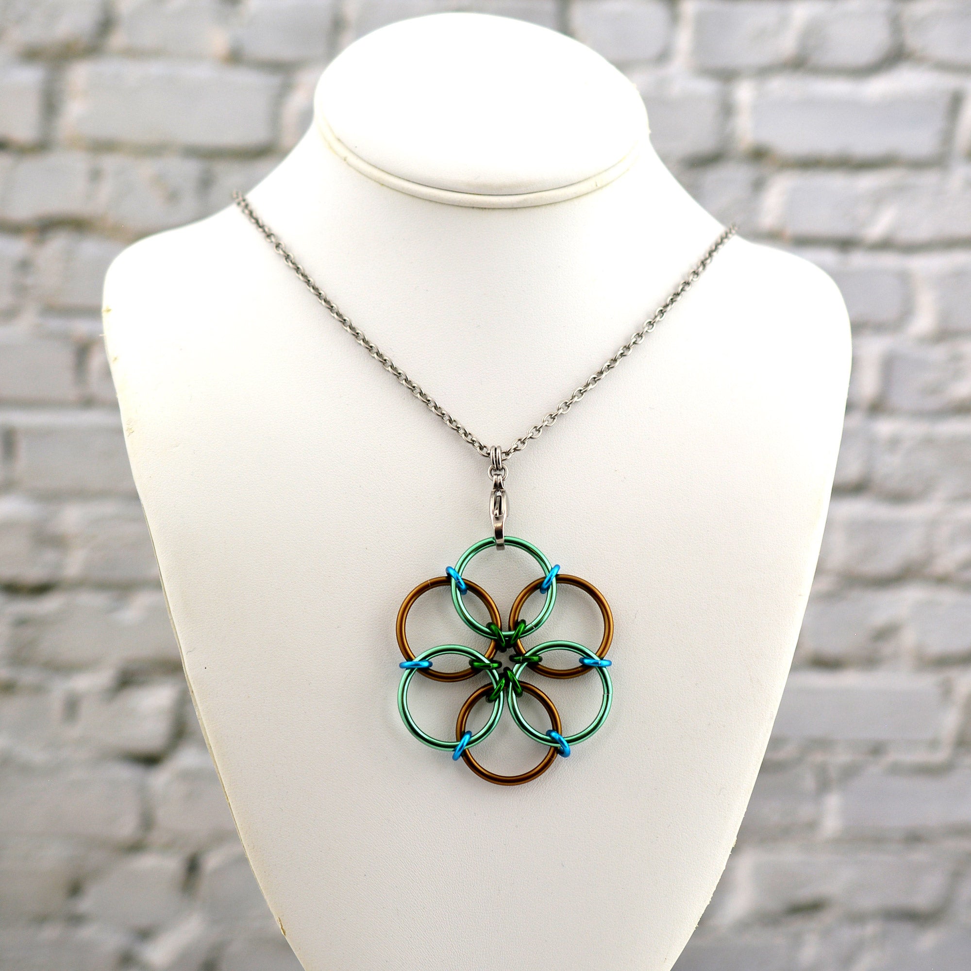 The Magical Woodland Fidget Flower necklace displayed on a white jewelry neck form against a white brick background. The pendant features mint green and russet brown rings with forest green and turquoise accents.