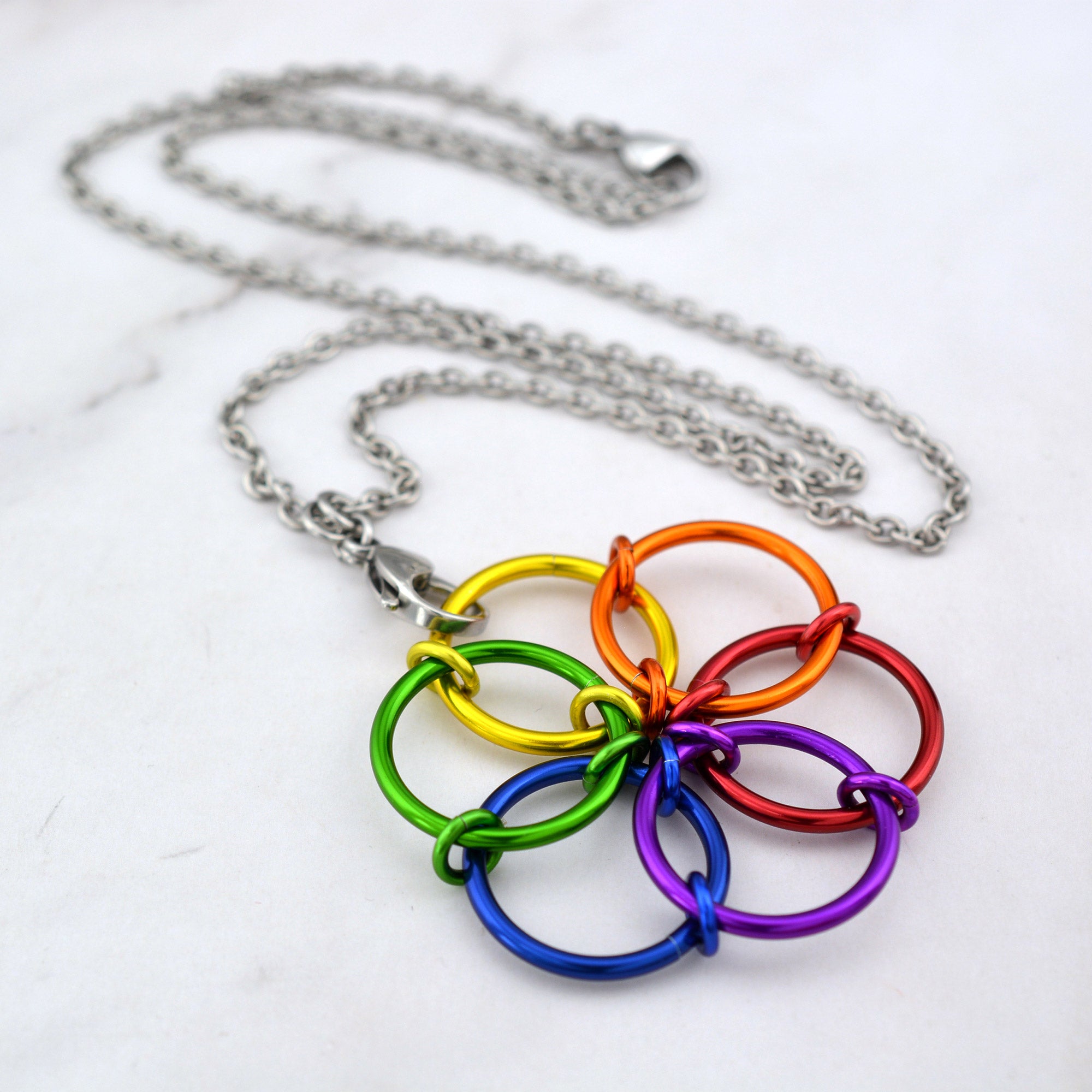 An angled aerial view of the Rainbow Fidget Flower necklace lying on a white marble surface, showing the vibrant rings in the sequence of red, orange, yellow, green, blue, and violet.
