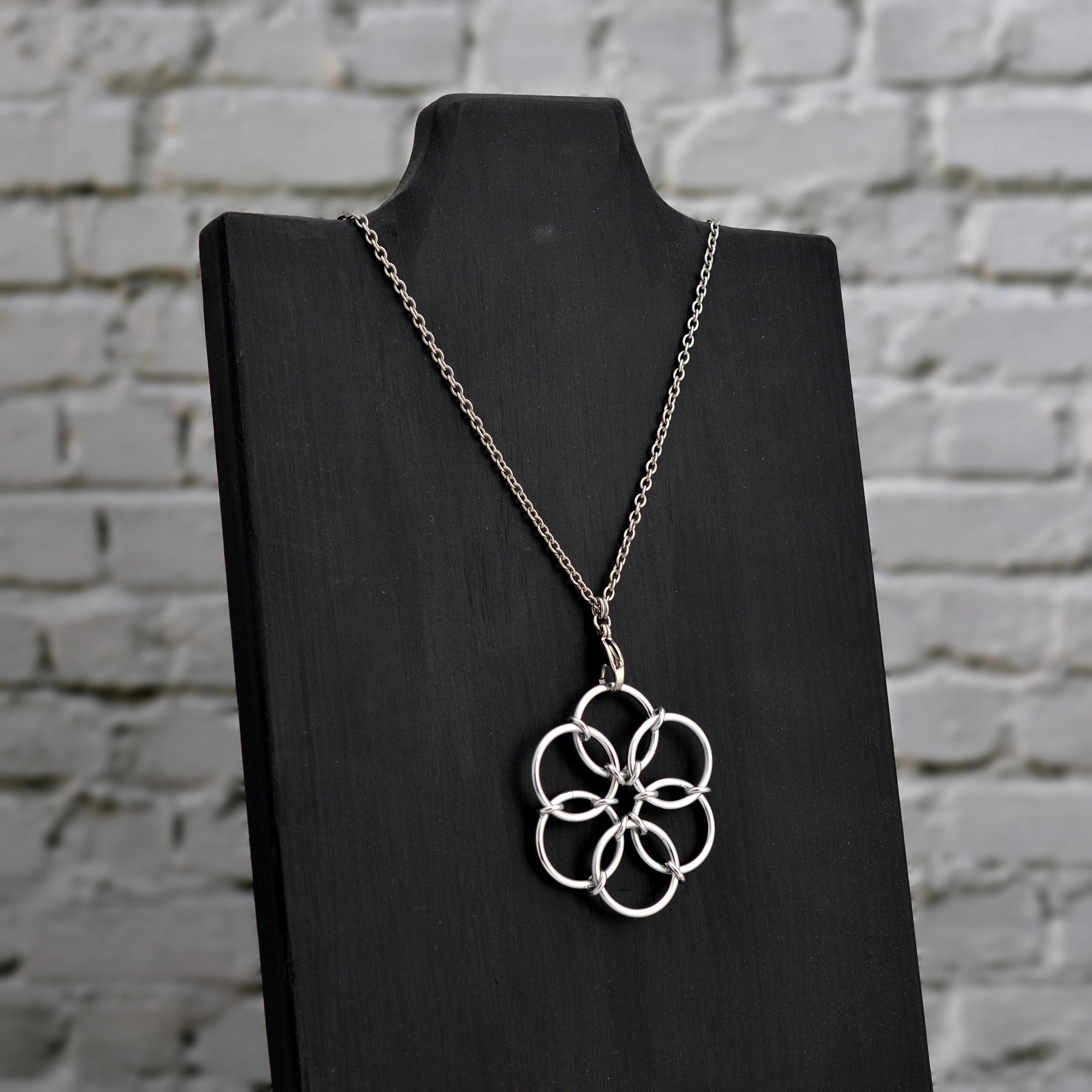 A silver Fidget Flower necklace displayed on a black jewelry stand, angled to the side, against a white brick wall background.